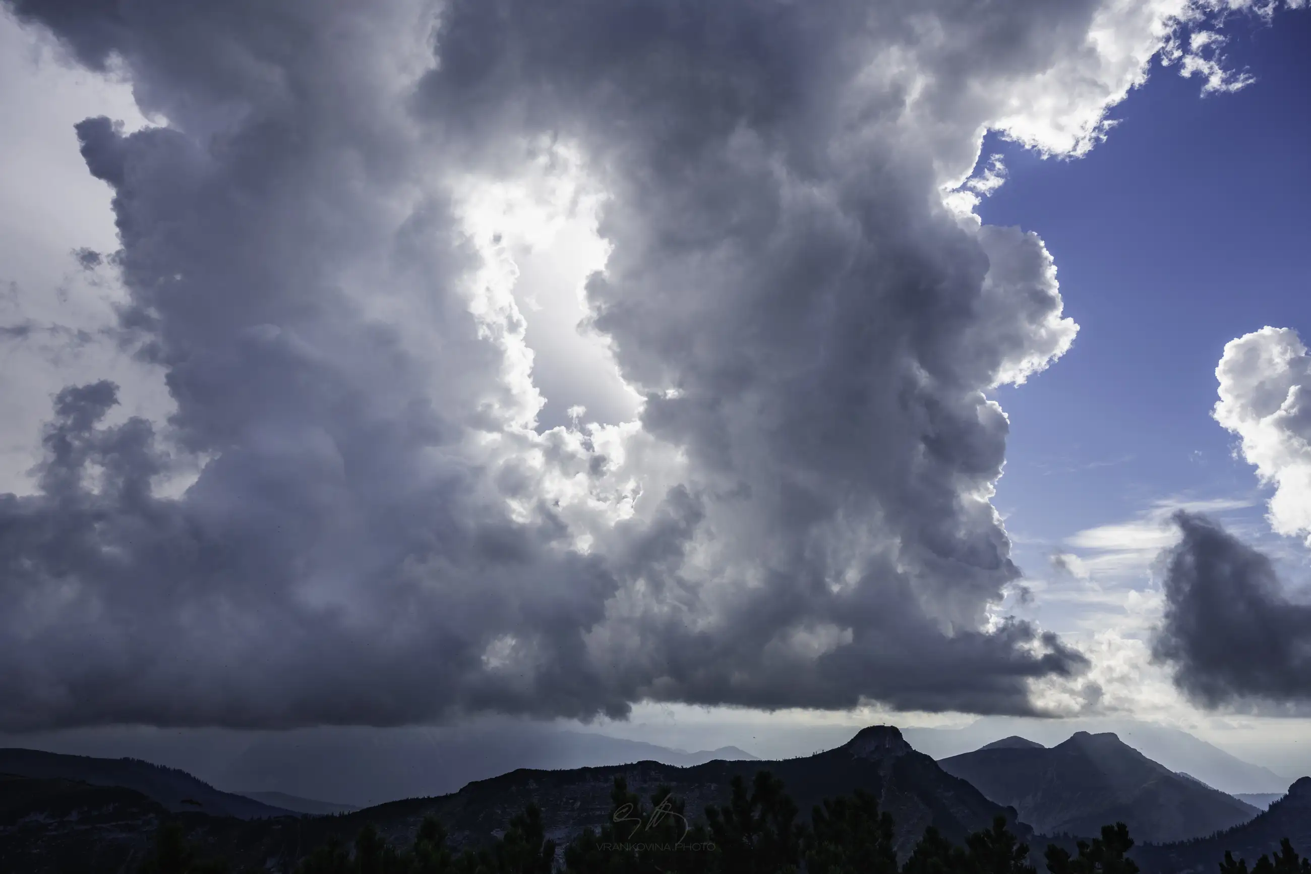 Large, dramatic clouds frame a bright blue sky above distant, shadowy mountain peaks, while dark green trees fill the foreground. Sunlight filters through the clouds, creating a dynamic and moody landscape scene.