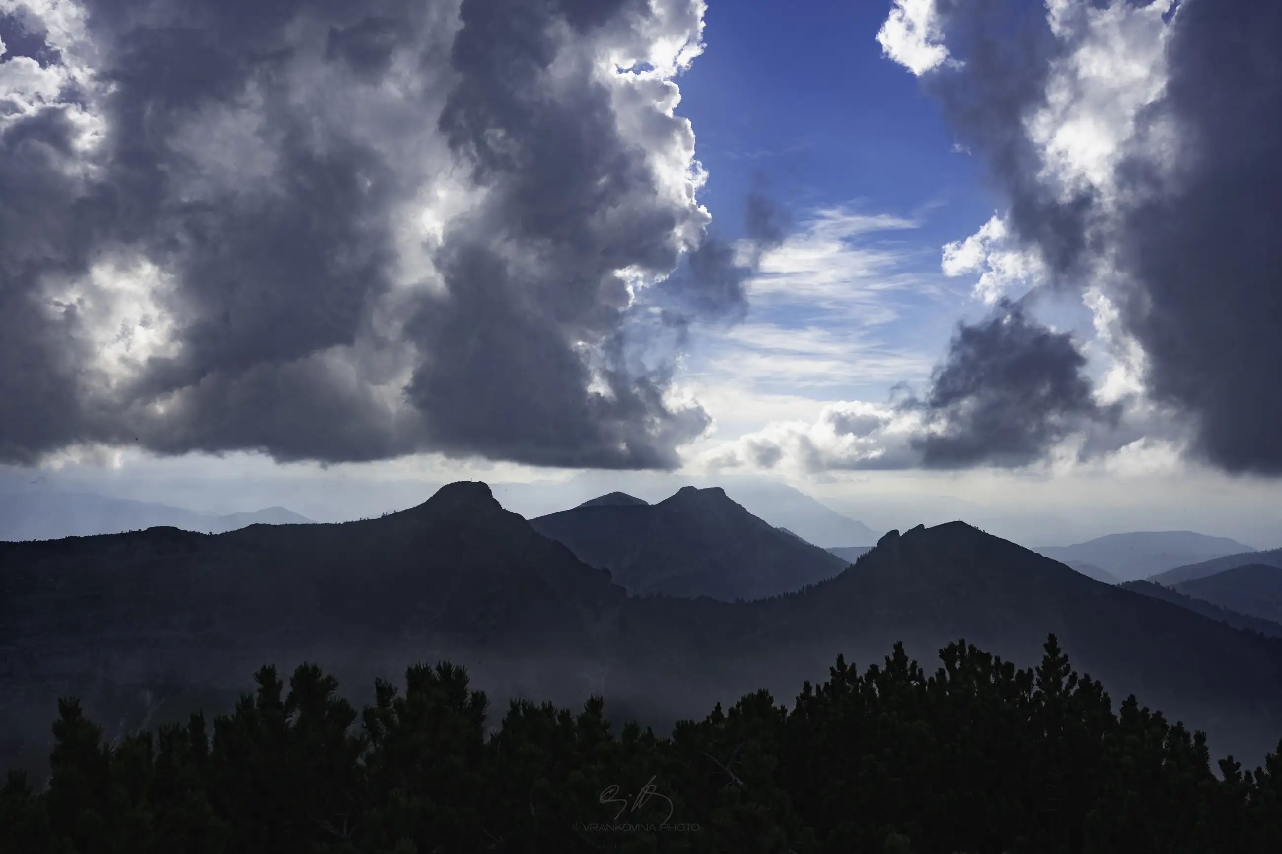 Large, dramatic clouds frame a bright blue sky above distant, shadowy mountain peaks, while dark green trees fill the foreground. Sunlight filters through the clouds, creating a dynamic and moody landscape scene.