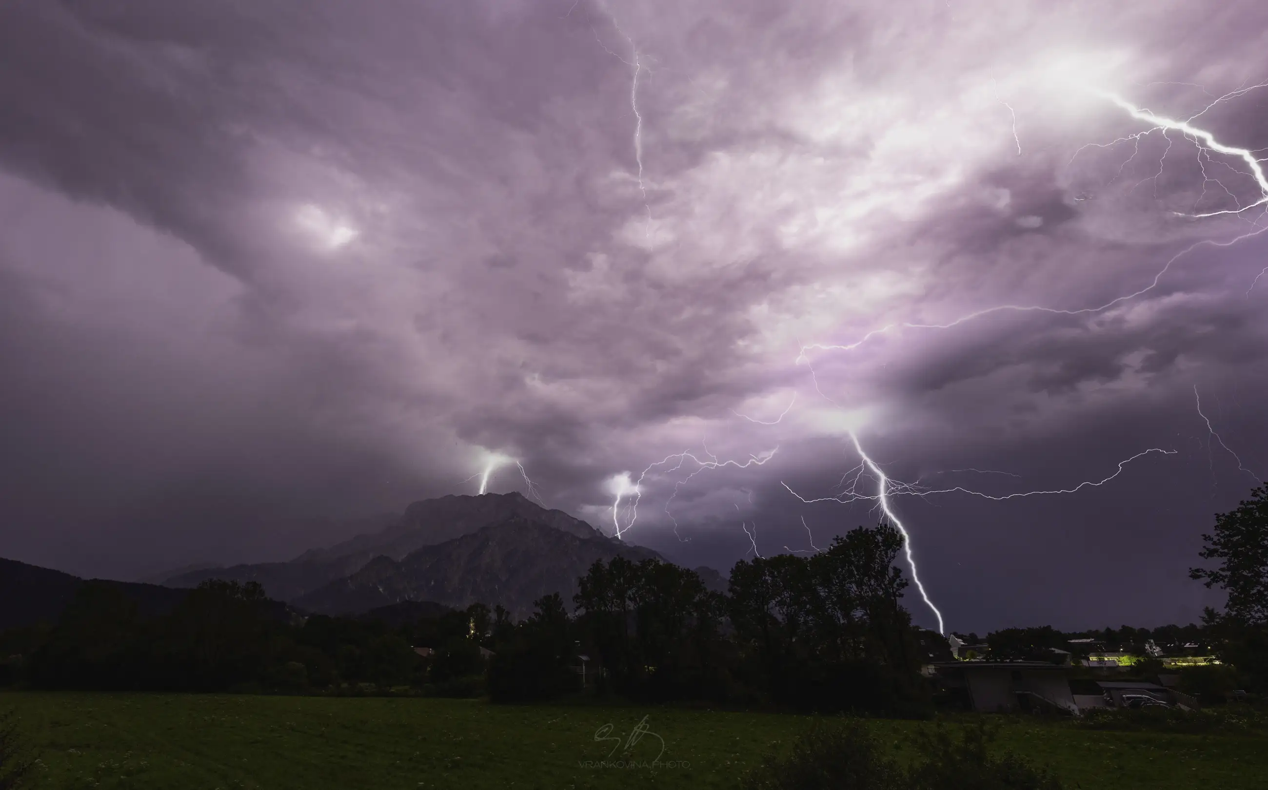 Lightning bolts strike down from dramatic, purple-tinged storm clouds over a mountain and a small town, illuminating the landscape and trees below during a nighttime thunderstorm.