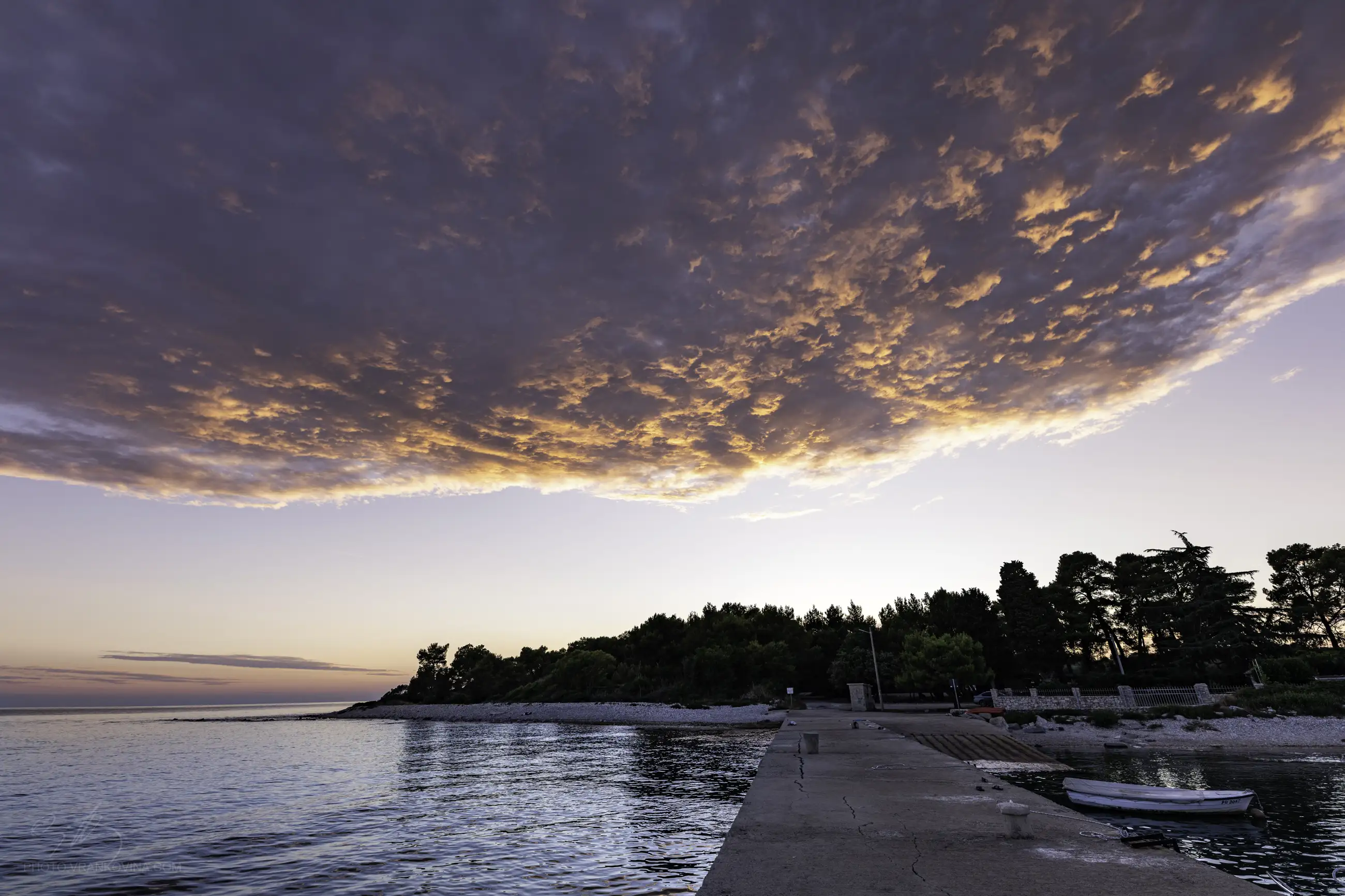 A concrete pier extends into calm water at sunset, with dramatic orange and purple clouds in the sky and a tree-lined shoreline in the background.