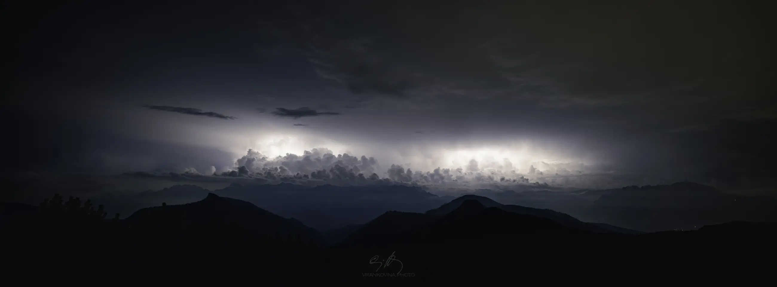 A dramatic night sky filled with dense clouds is illuminated by flashes of lightning above dark mountain silhouettes. The horizon glows with white and orange hues, highlighting the intense storm.