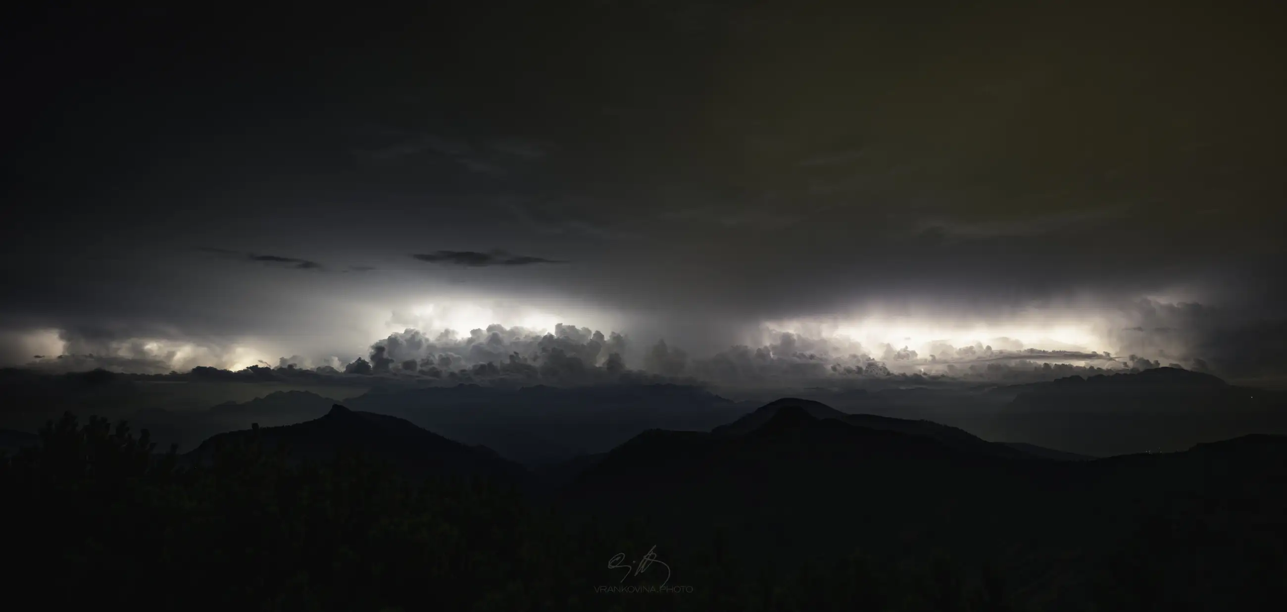 A dramatic night sky filled with dense clouds is illuminated by flashes of lightning above dark mountain silhouettes. The horizon glows with white and orange hues, highlighting the intense storm.