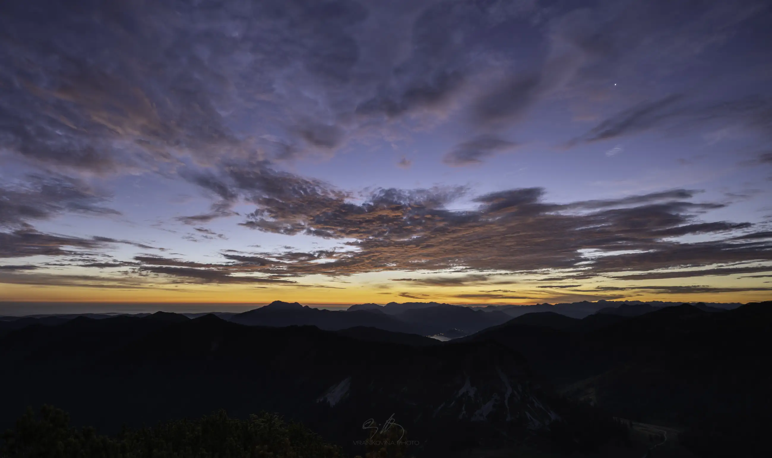 A dramatic sunset over mountain ranges, with dark silhouettes of peaks and a golden-orange sky partly covered by textured clouds. A lake is visible in the distant valley below.