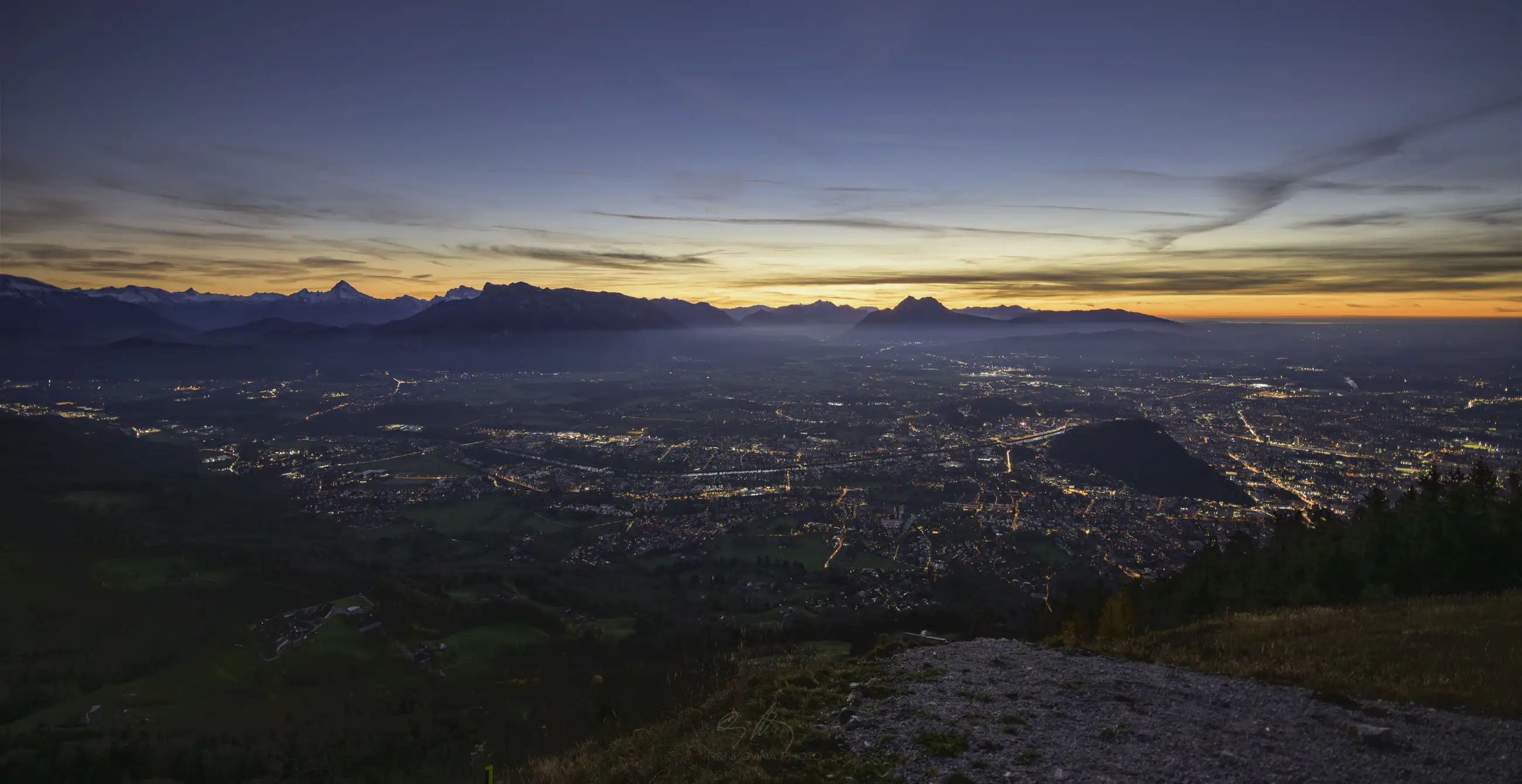 A scenic view of a city at dusk, with twinkling lights spread across the valley, surrounded by mountains under a darkening sky with a glowing sunset near the horizon.