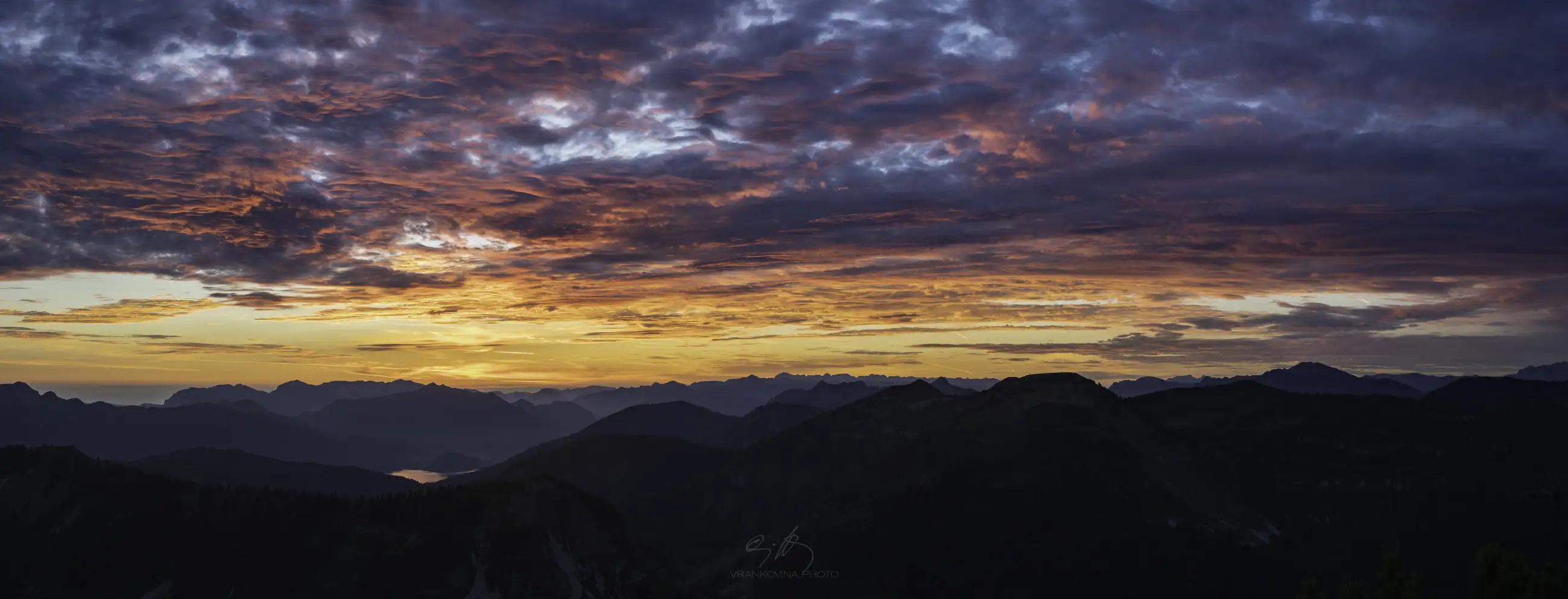 A vibrant sunset over mountain ranges, with the sun low on the horizon casting orange and purple hues across dramatic, cloud-filled skies and silhouetted peaks in the foreground.