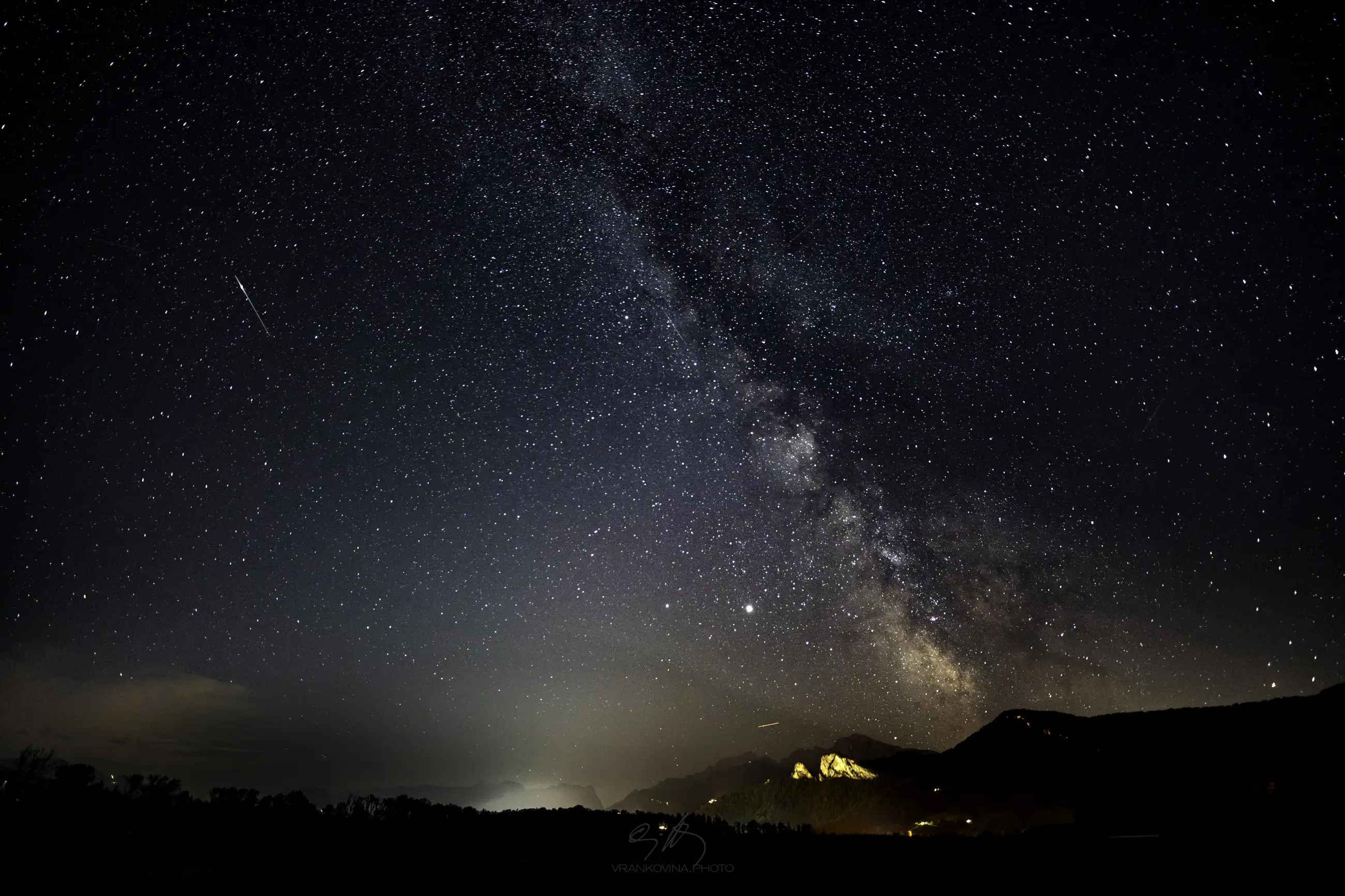 A clear night sky filled with stars and a shooting star, the bright band of the Milky Way stretching diagonally above dark silhouetted mountains and a softly lit horizon.