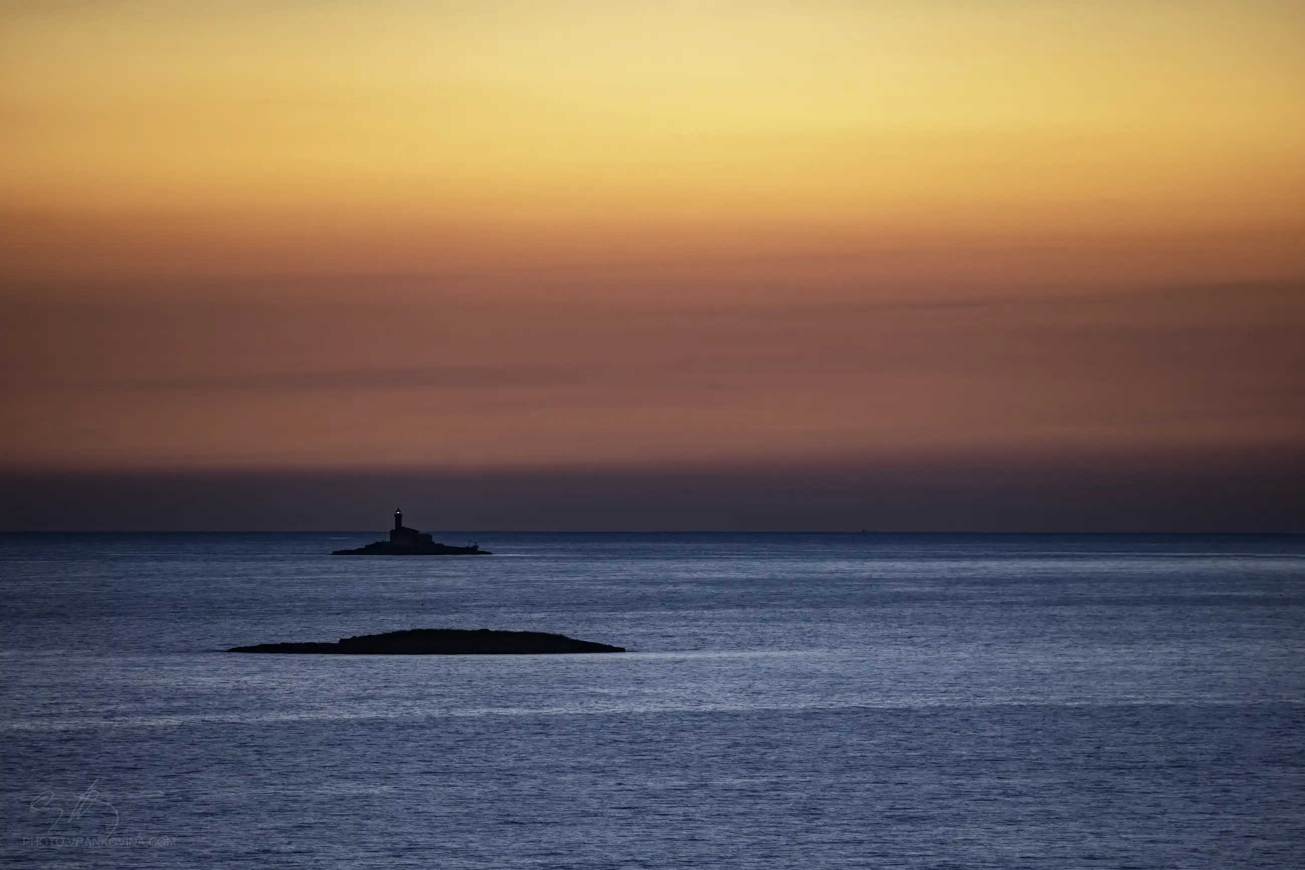 A small lighthouse stands on a distant island under a colorful sunset sky, with calm blue water and another smaller, rocky island in the foreground.