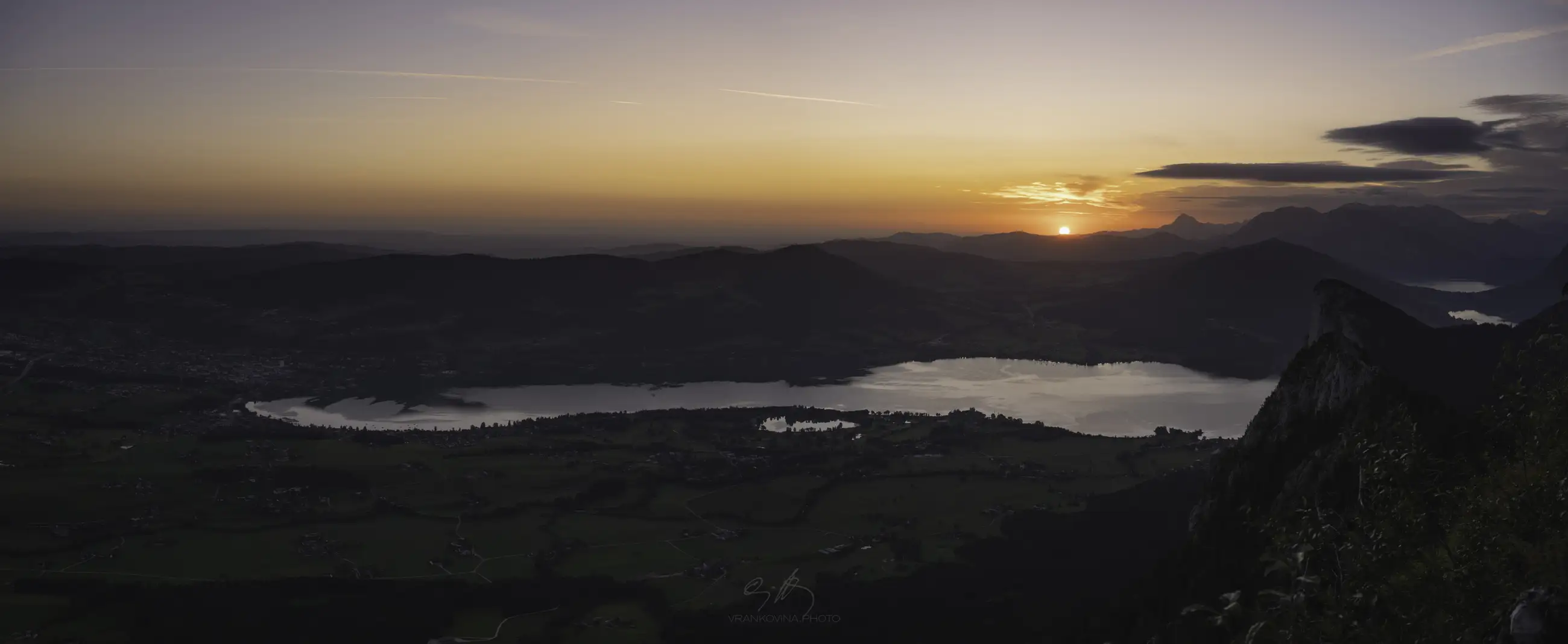 A panoramic view of a lake surrounded by hills and mountains at sunset, with the sun setting behind distant peaks and the sky glowing with warm colors.