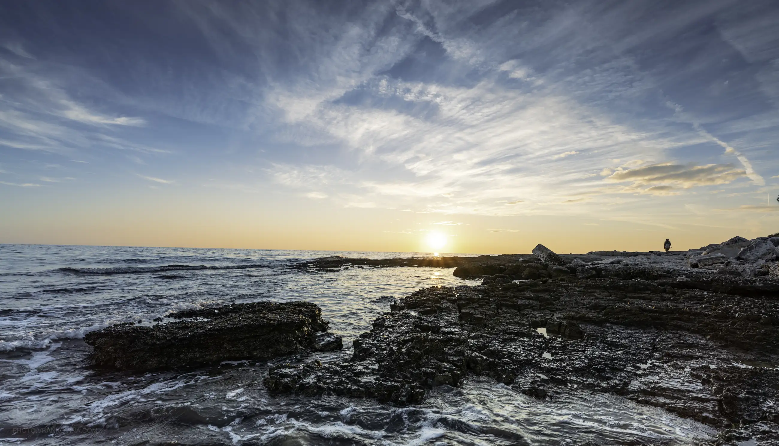 Rocky shoreline with gentle waves under a blue sky, as the sun sets on the horizon. Wispy clouds are scattered across the sky and a lone figure stands on the rocks in the distance.