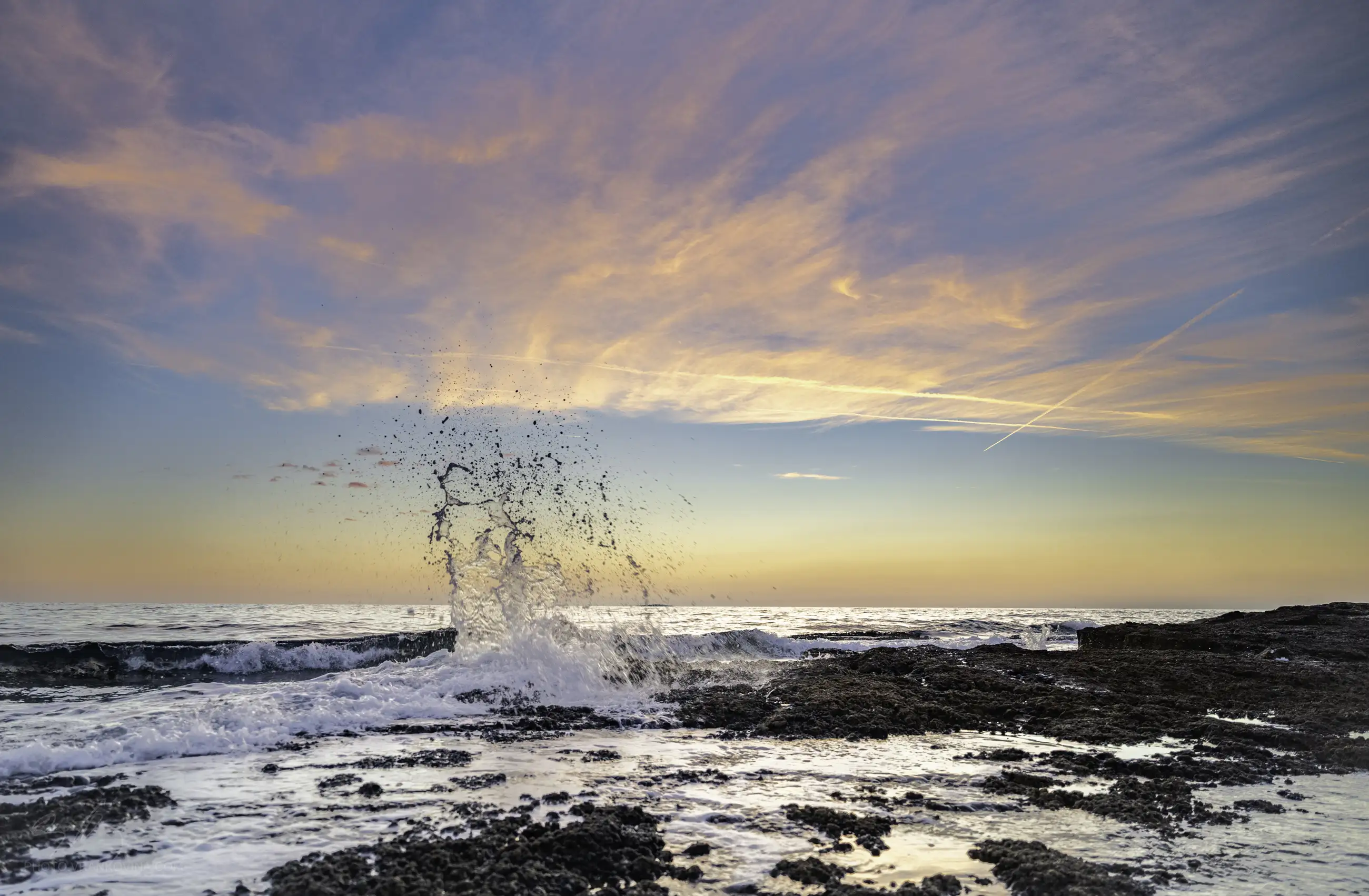 Waves crash against rocky shore at sunset, sending water spray into the air. The sky is painted with pink and orange clouds, and the calm ocean stretches to the horizon.