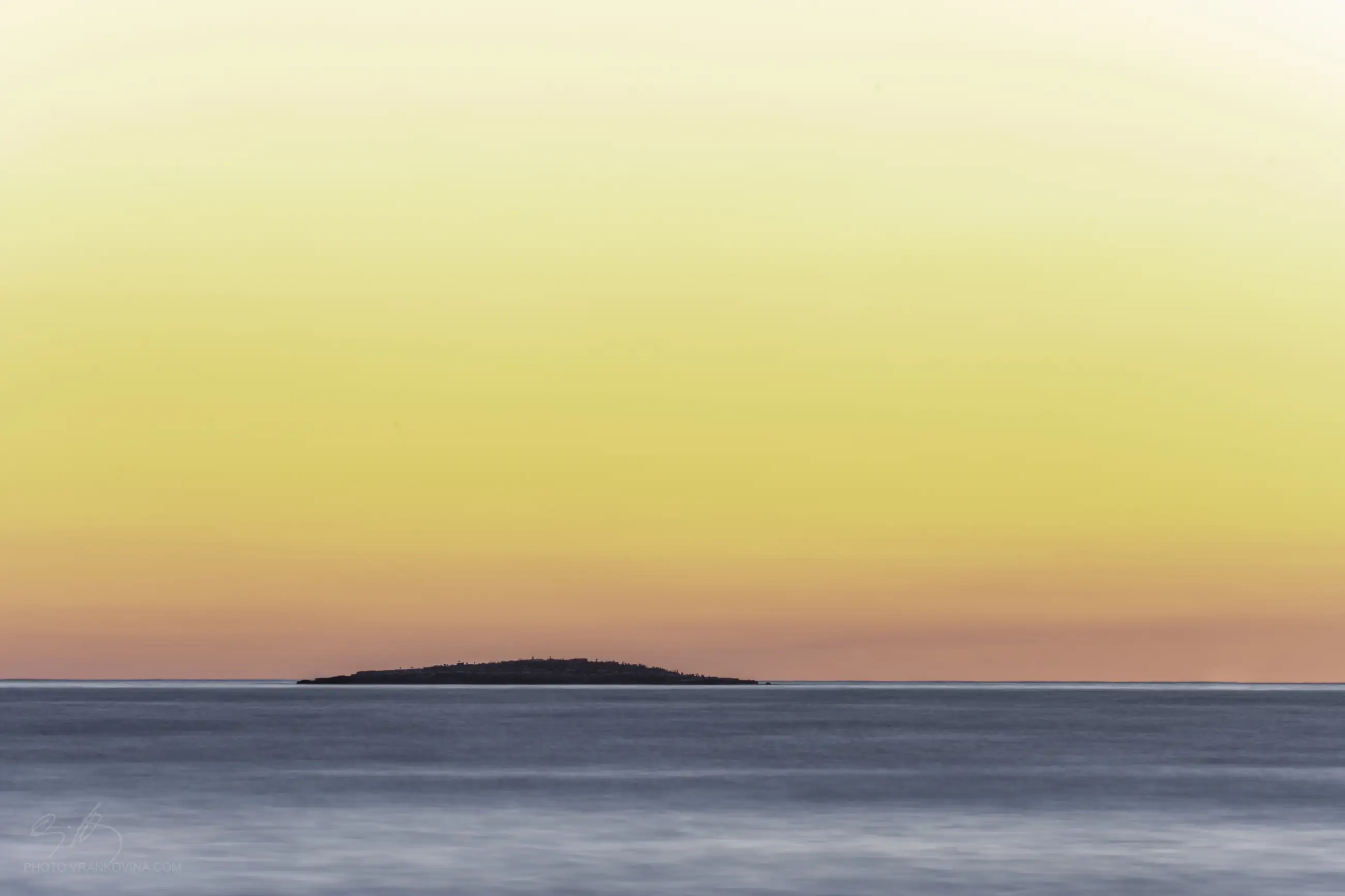 A distant, low island sits on a calm ocean beneath a gradient sky of yellow, orange, and pink at sunrise or sunset, creating a serene and minimalist seascape.