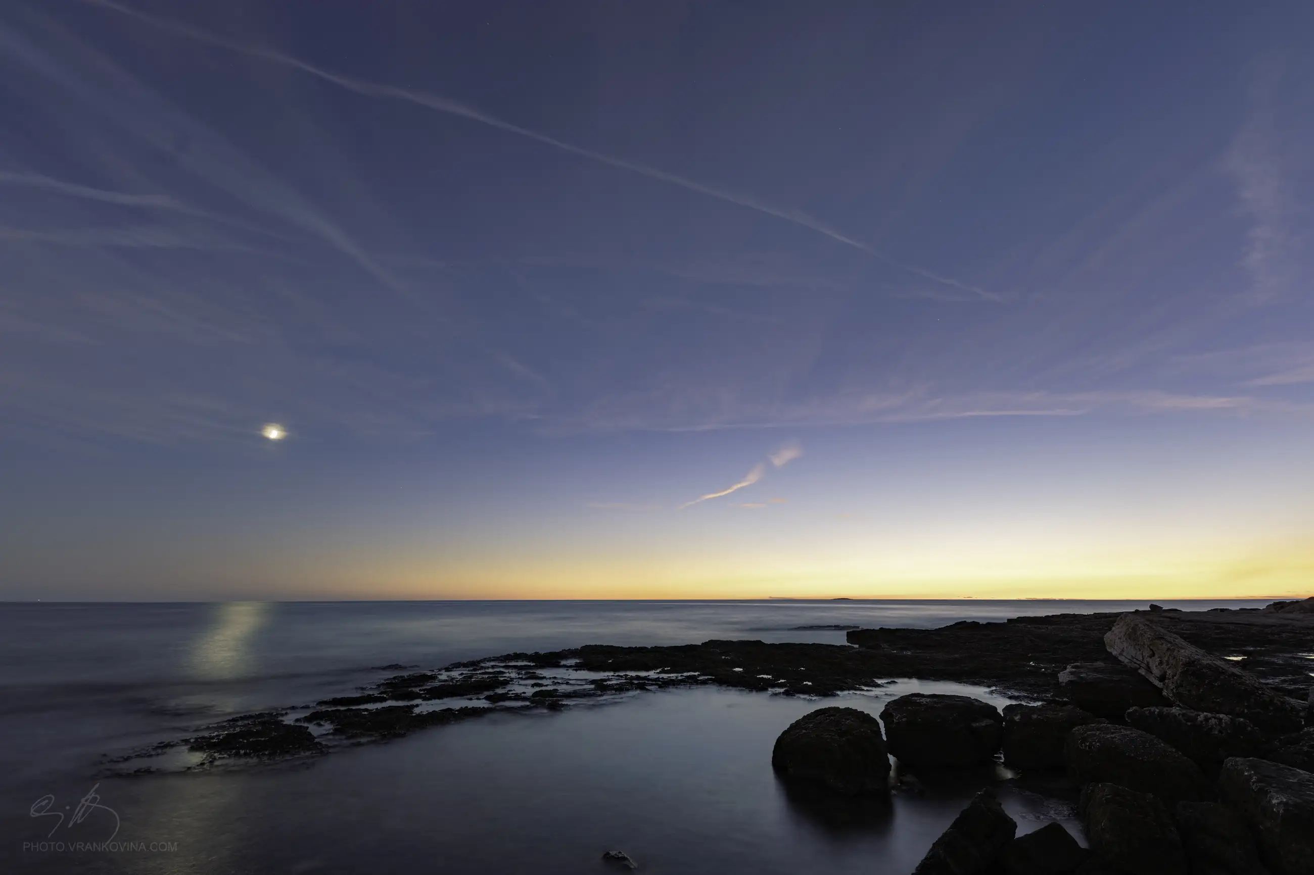 A calm coastal scene at dusk, with a glowing sunset on the horizon, a crescent moon in the sky, and gentle waves lapping against dark rocks along the shoreline. Contrails are faintly visible in the sky.