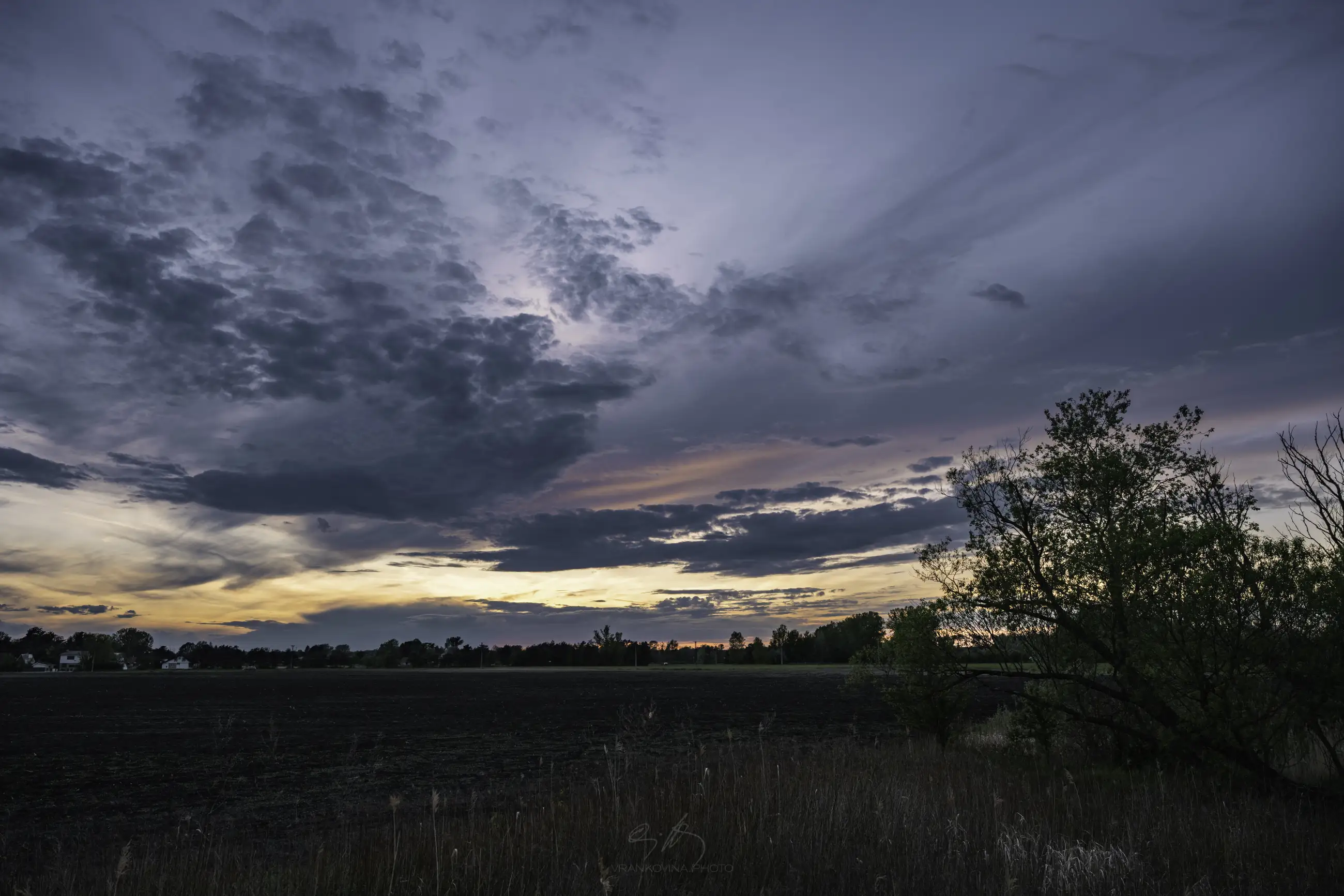 A wide, open field at sunset with dramatic, dark clouds streaking across the sky. The horizon glows orange and pink, while trees and grass frame the landscape in silhouette.