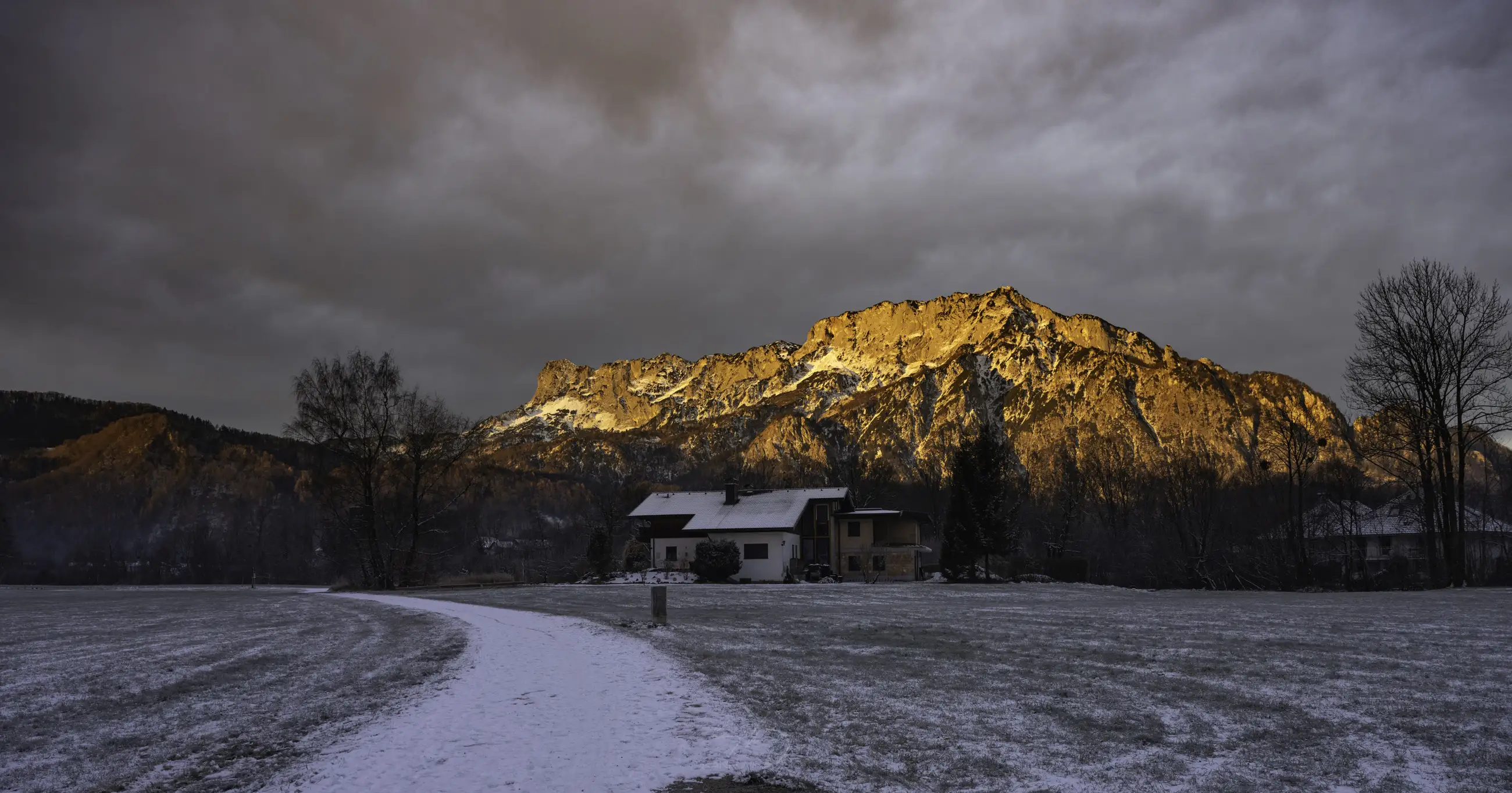 A small house sits in a snowy field under dramatic, cloudy skies, with a mountain in the background glowing orange in the light of the setting or rising sun.