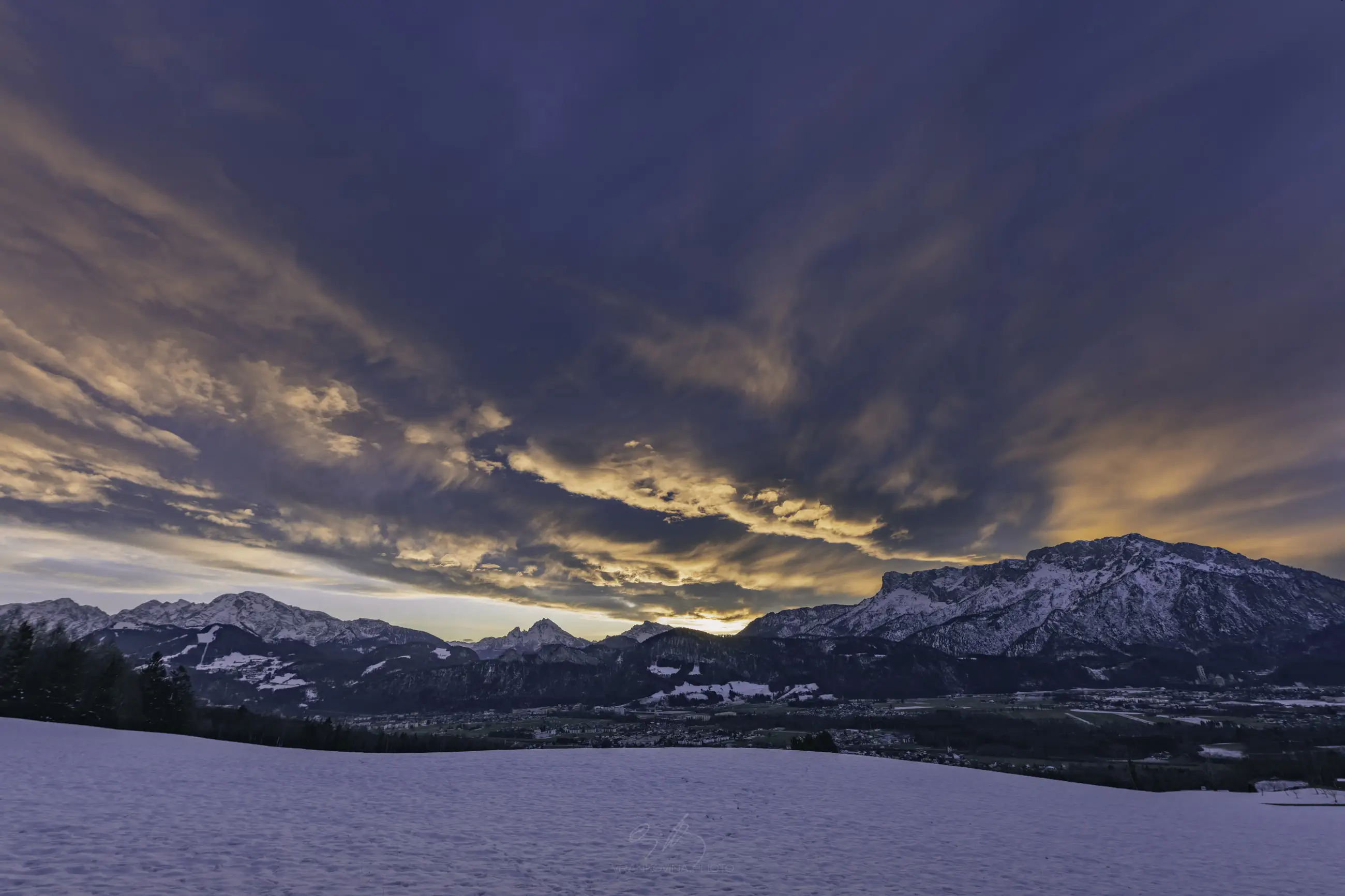 Snow-covered field with distant mountains under a dramatic sky filled with orange and purple clouds at sunset. The scene is peaceful, with the sun partially hidden behind the peaks.