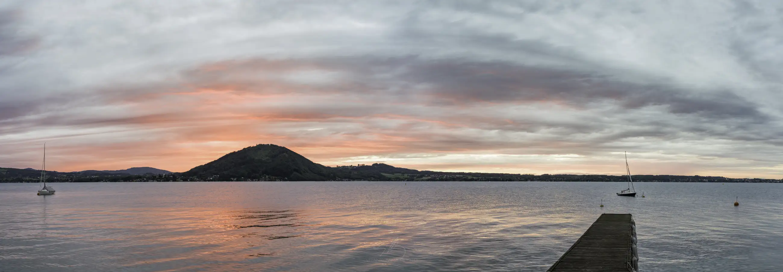 A calm lake at sunset with pink and orange clouds, a mountain in the background, two sailboats on the water, and a wooden dock extending into the lake from the right.