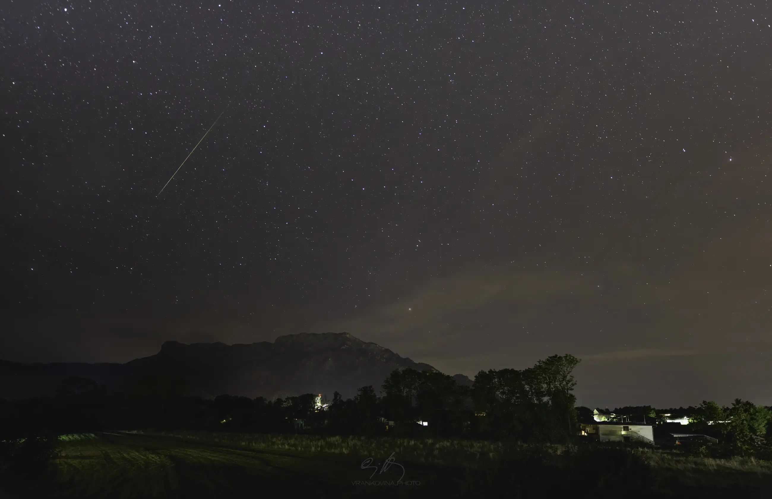 A night sky filled with stars and a shooting star above a mountain range. Below, lights from houses illuminate the landscape, with trees and buildings visible in the foreground.