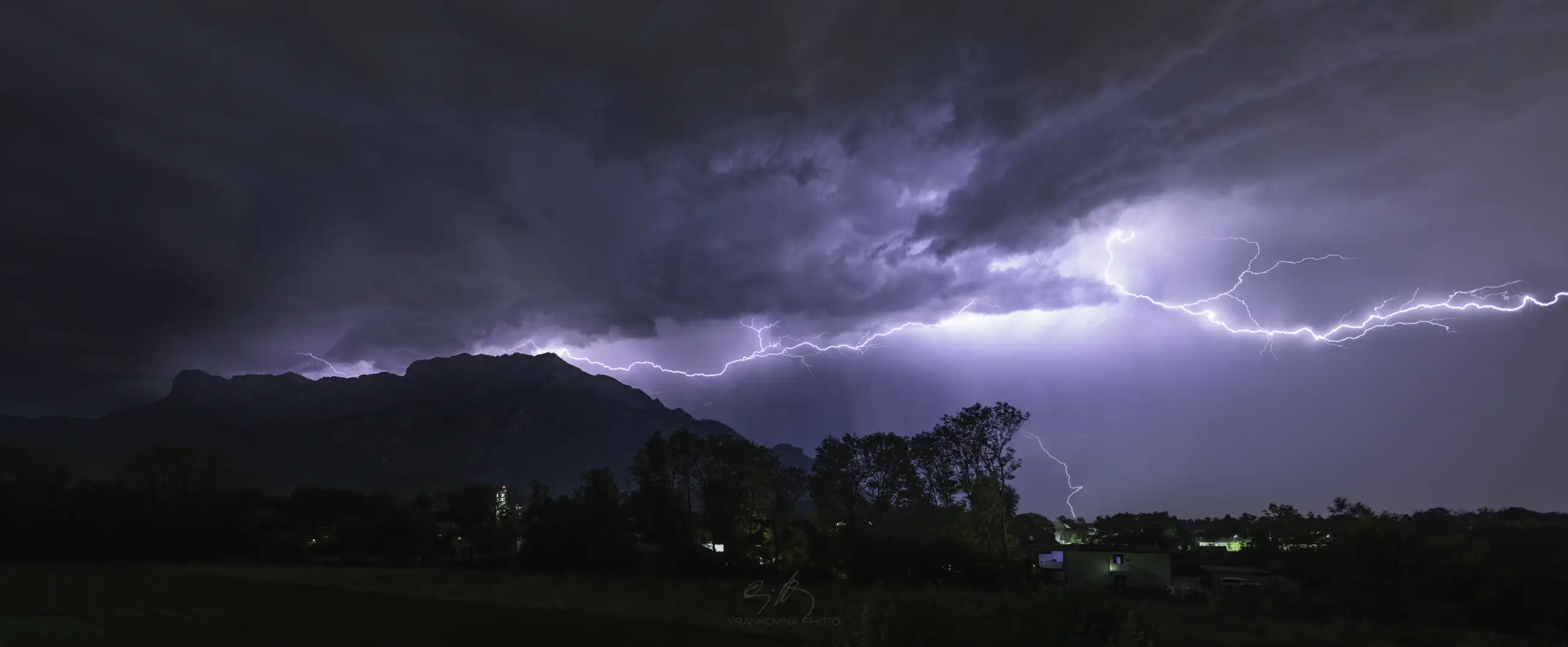 A dramatic night scene shows bright lightning illuminating dark storm clouds above a silhouette of mountains and trees, with faint lights from houses below.