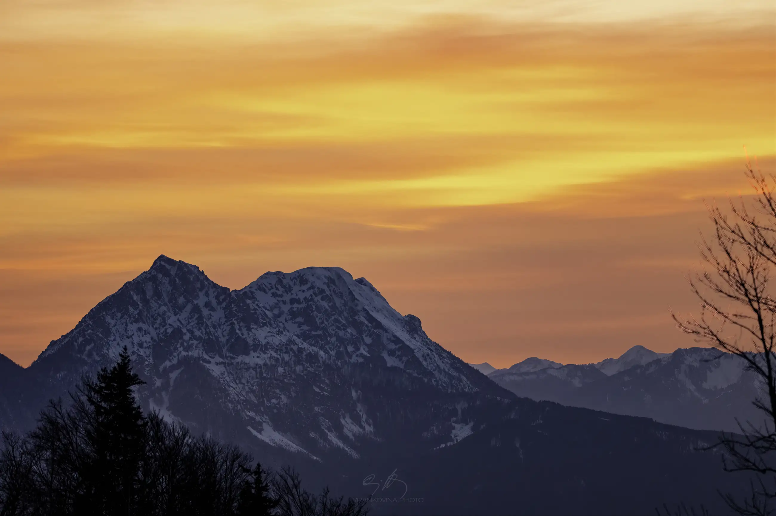 Snow-capped mountain silhouettes under a vibrant sunset sky with layers of orange, pink, and blue clouds. Pine tree branches frame the scene on the right side.