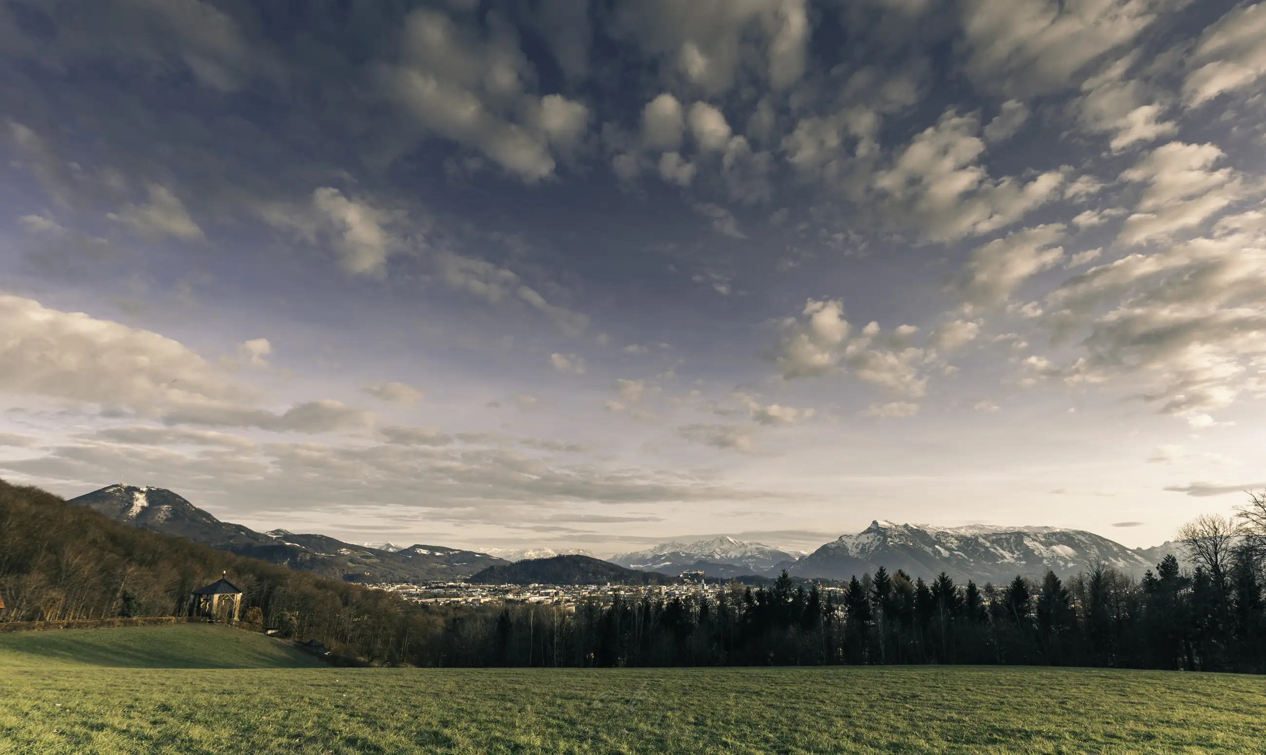 A scenic landscape with a grassy field in the foreground, trees and hills in the middle, distant snow-capped mountains, and a town nestled below a sky with scattered clouds.