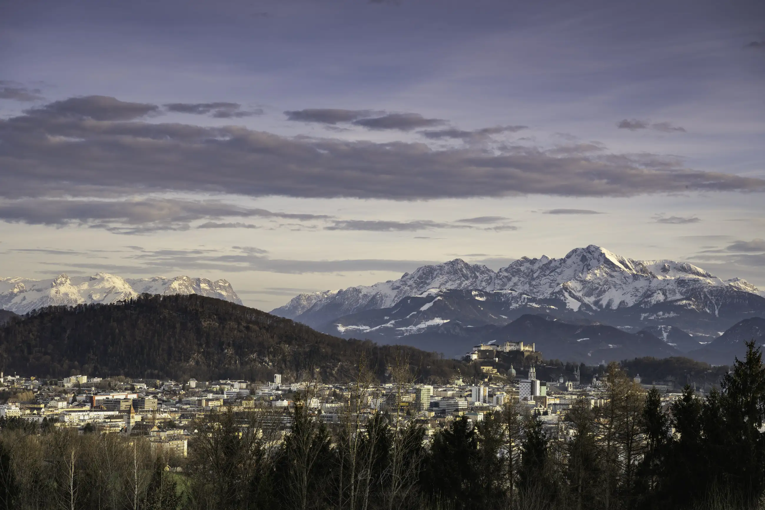 A scenic view of a city nestled among trees with snow-capped mountains in the background under a cloudy sky at dusk.