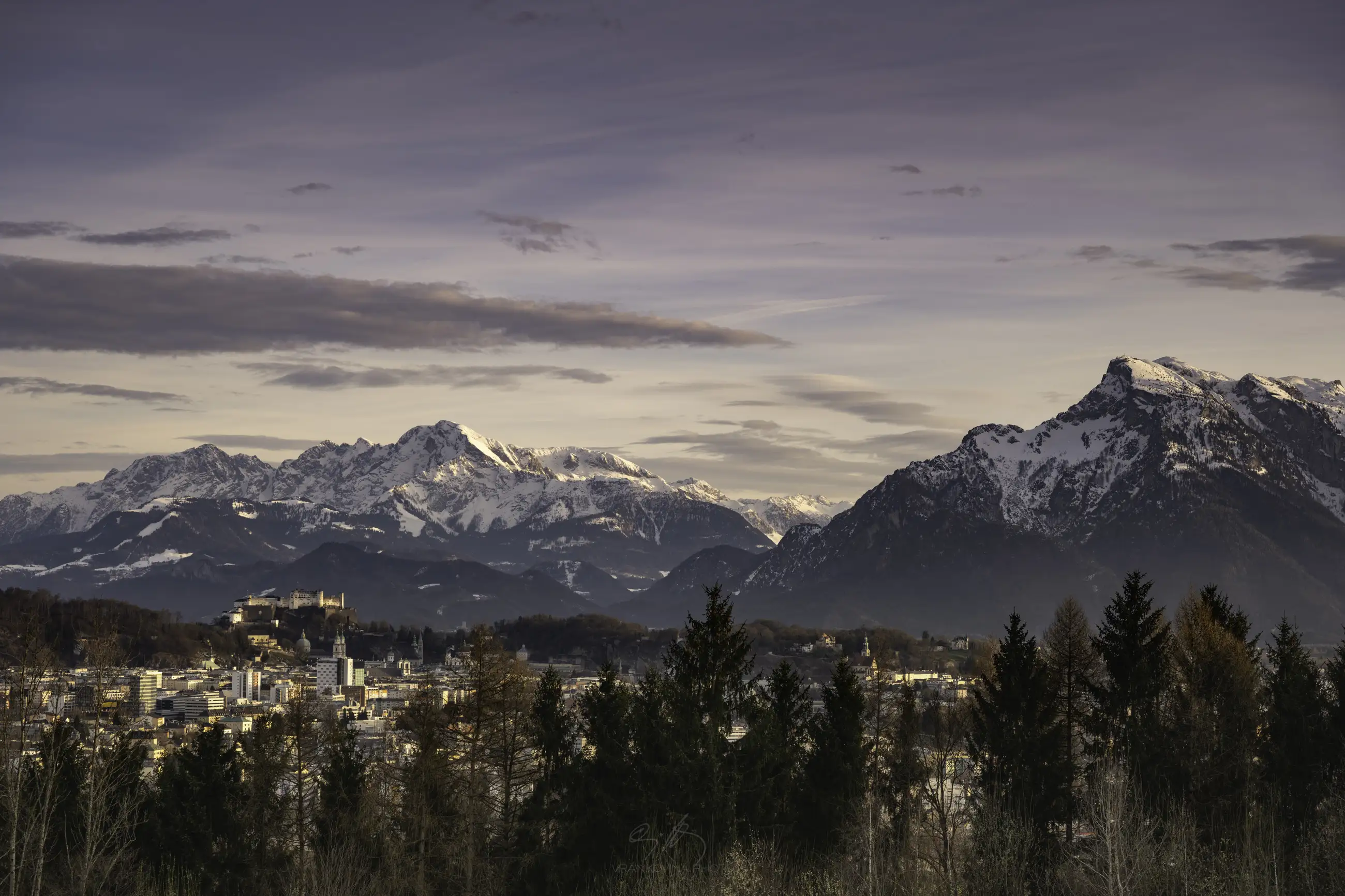 A scenic view of a city nestled among trees with snow-capped mountains in the background under a cloudy sky at dusk.