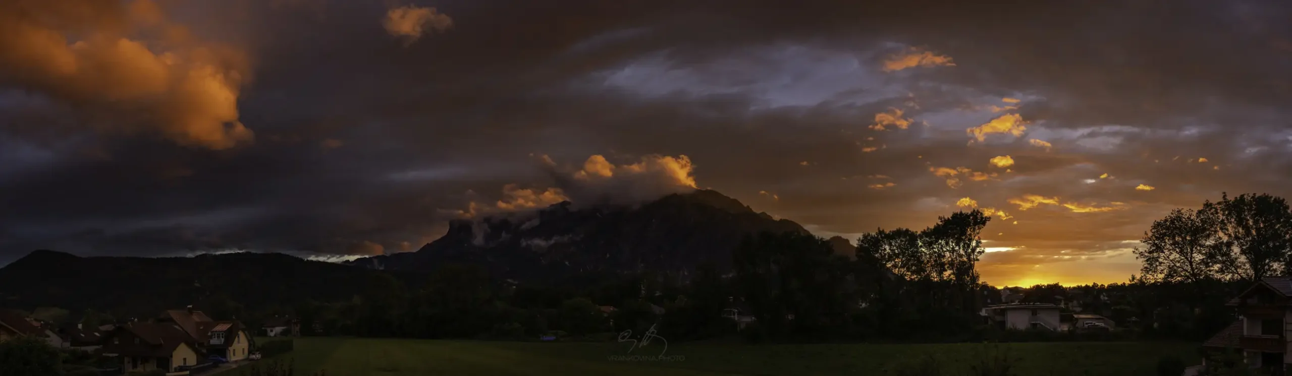 A panoramic view, dramatic sunset with orange and pink clouds illuminates a mountain partially covered in clouds, with trees and houses in silhouette in the foreground.
