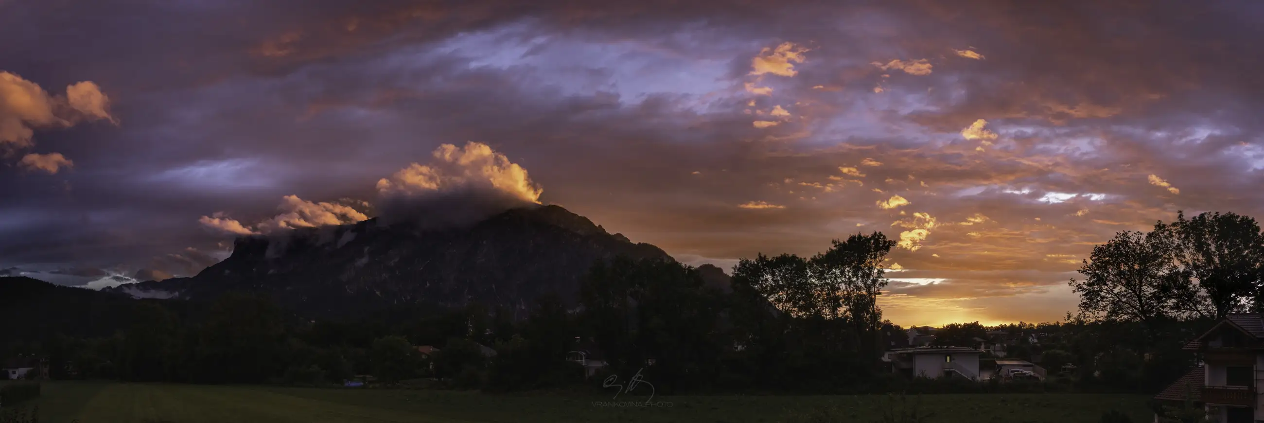A dramatic sunset with orange and pink clouds illuminates a mountain partially covered in clouds, with trees and houses in silhouette in the foreground.