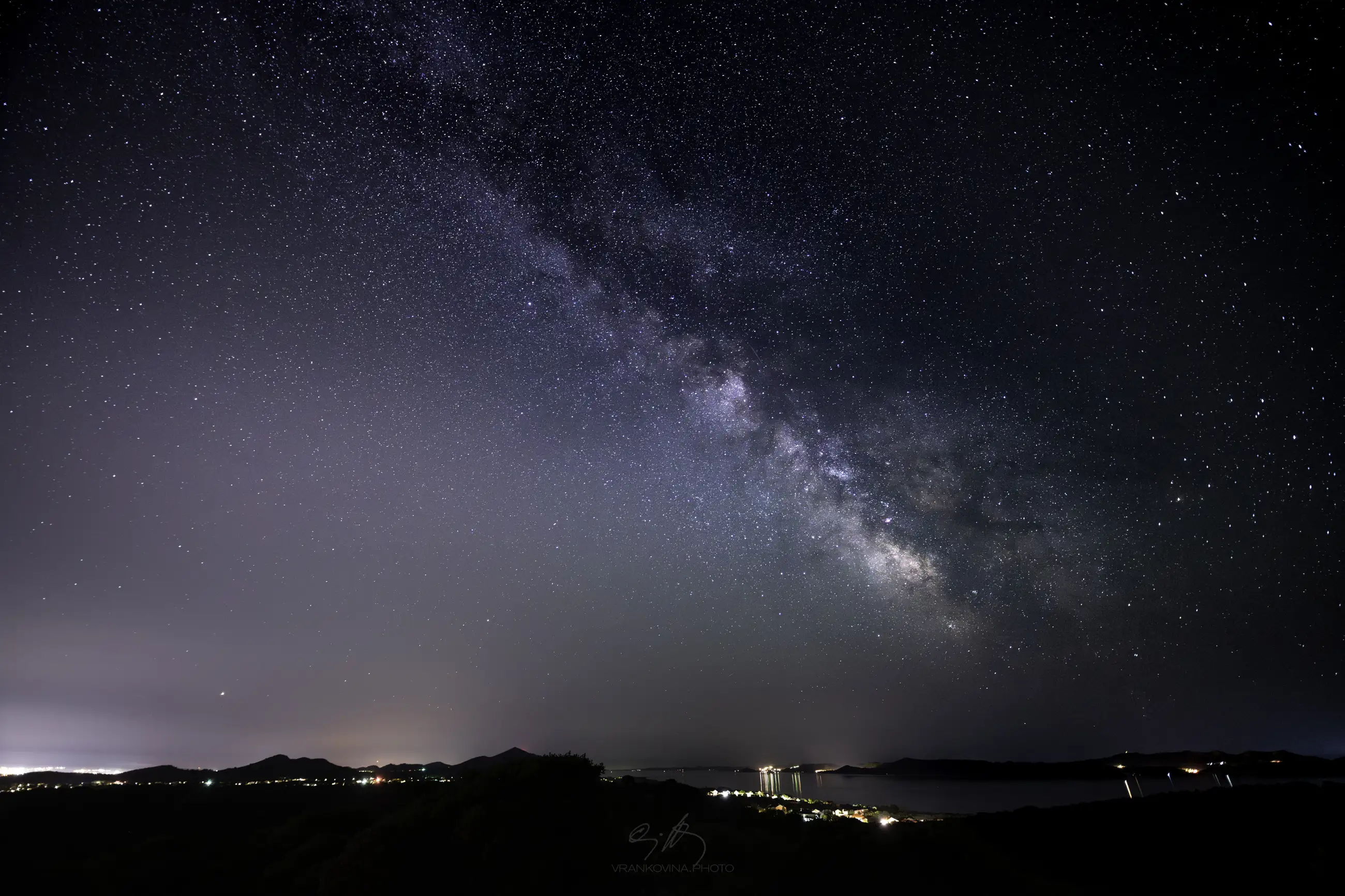 A clear night sky shows the Milky Way stretching above a calm landscape with distant hills and a sea visible between islands, while scattered lights from buildings and reflections are visible in the foreground.