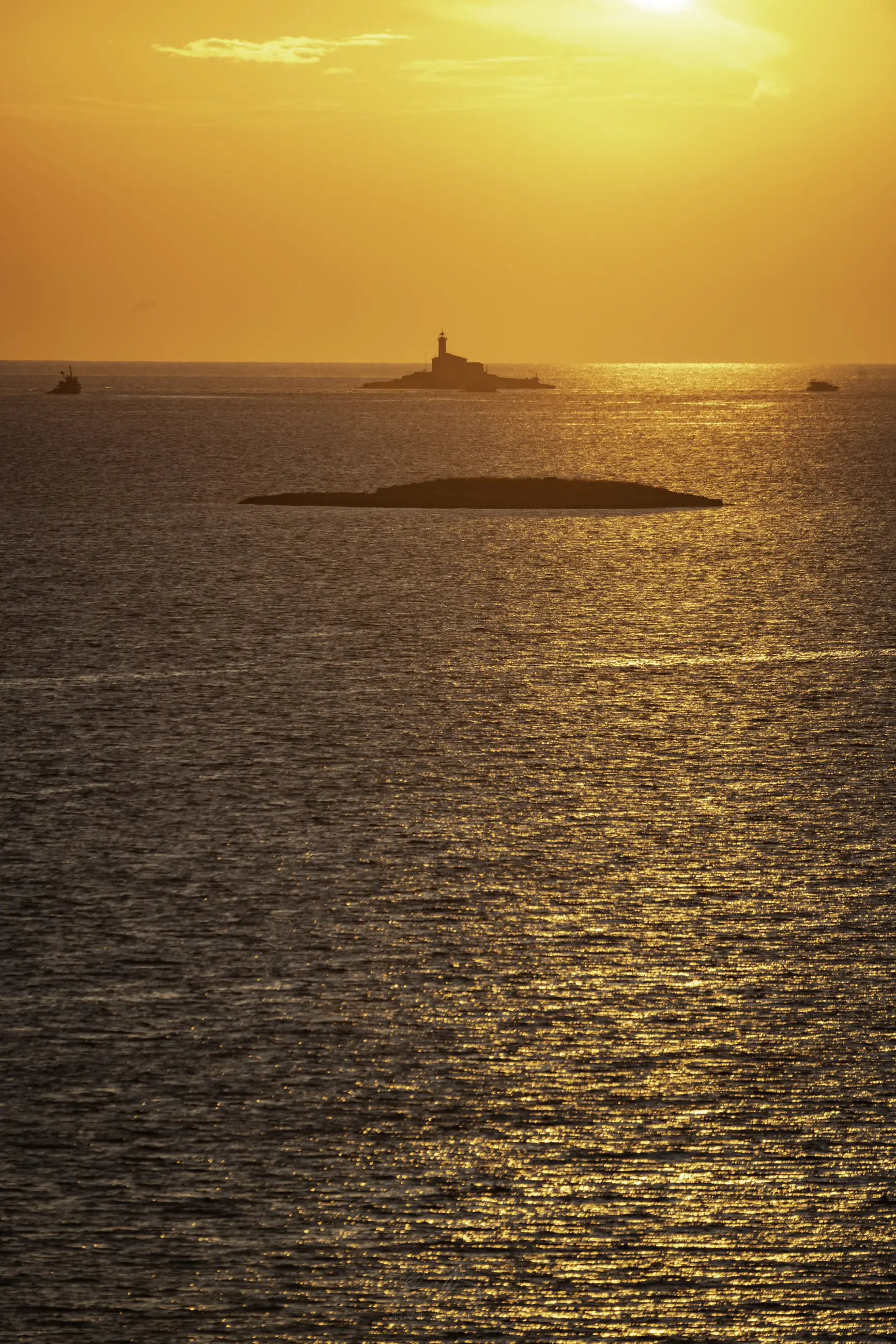 Golden sunset over a calm sea, with a lighthouse silhouetted on a distant small island. Several boats and rocky islets are visible on the shimmering water under the warm orange sky.