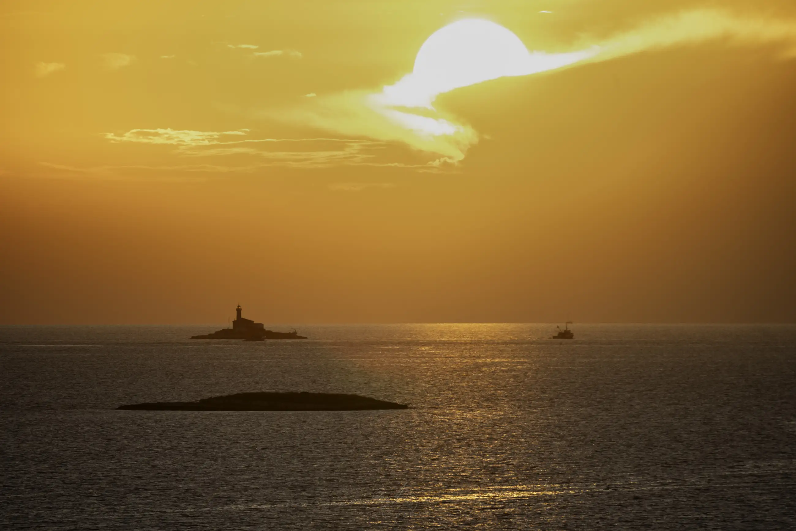 A vibrant orange sunset over the sea, with the sun partially covered by clouds. A small island with a lighthouse and another tiny island are visible, along with a boat on the calm water.
