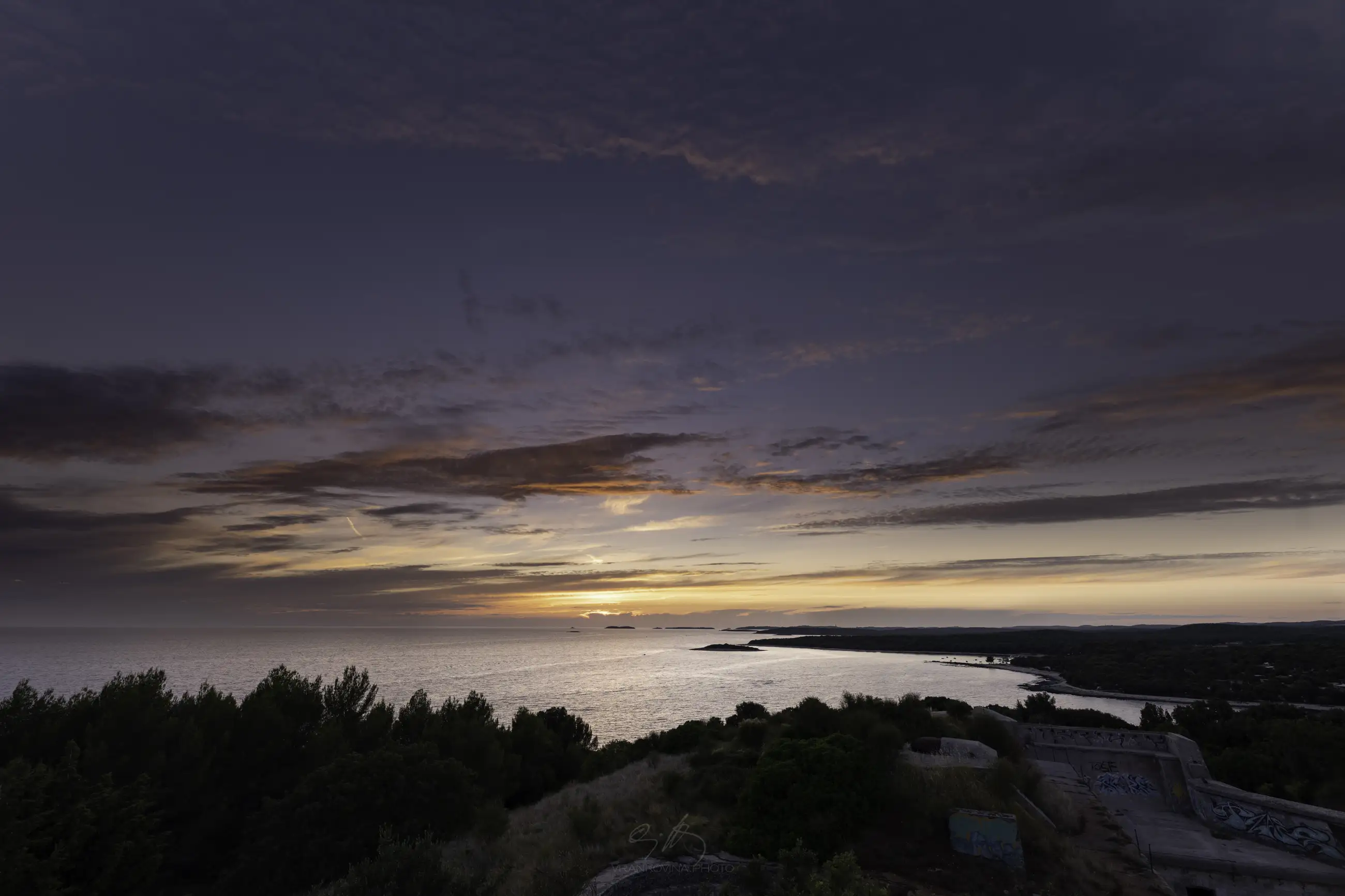 Sunset over a calm sea with dramatic clouds, seen from a coastal hill covered in green trees and shrubs. Some graffiti-covered structures are visible in the foreground.