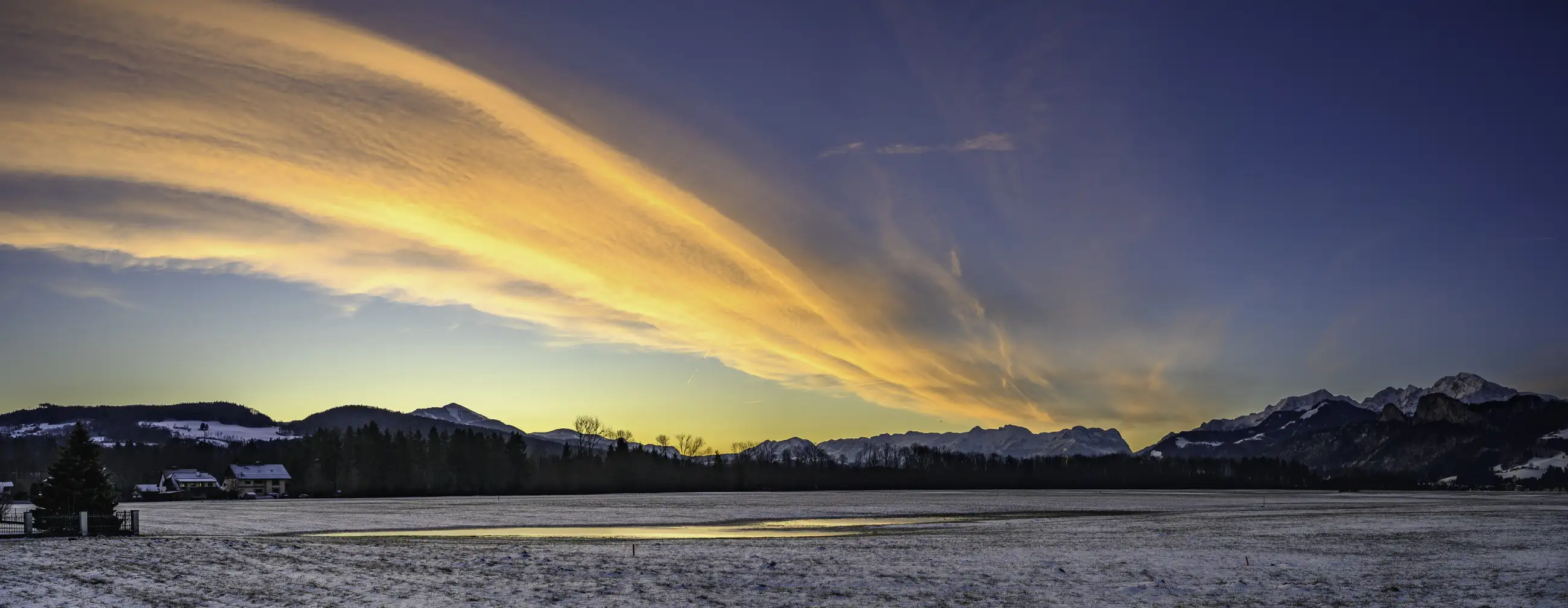 A panoramic view of a snowy landscape at sunrise, with vibrant orange clouds streaking across the sky above dark mountains and a few houses scattered near the tree line.