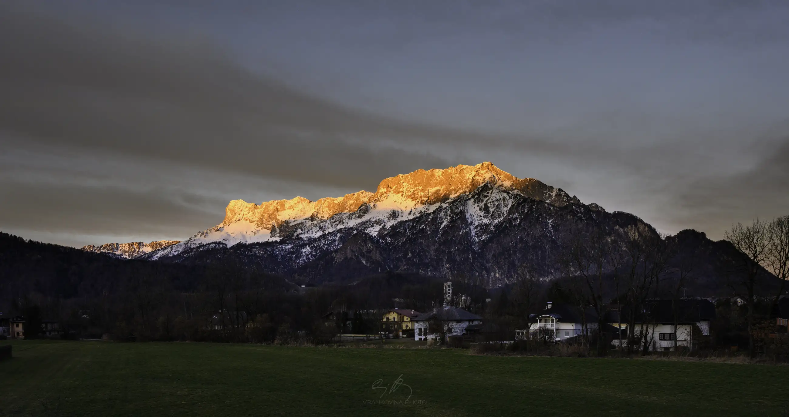 A mountain range with snow-capped peaks illuminated by the orange glow of sunrise, overlooking houses and green fields in the foreground.