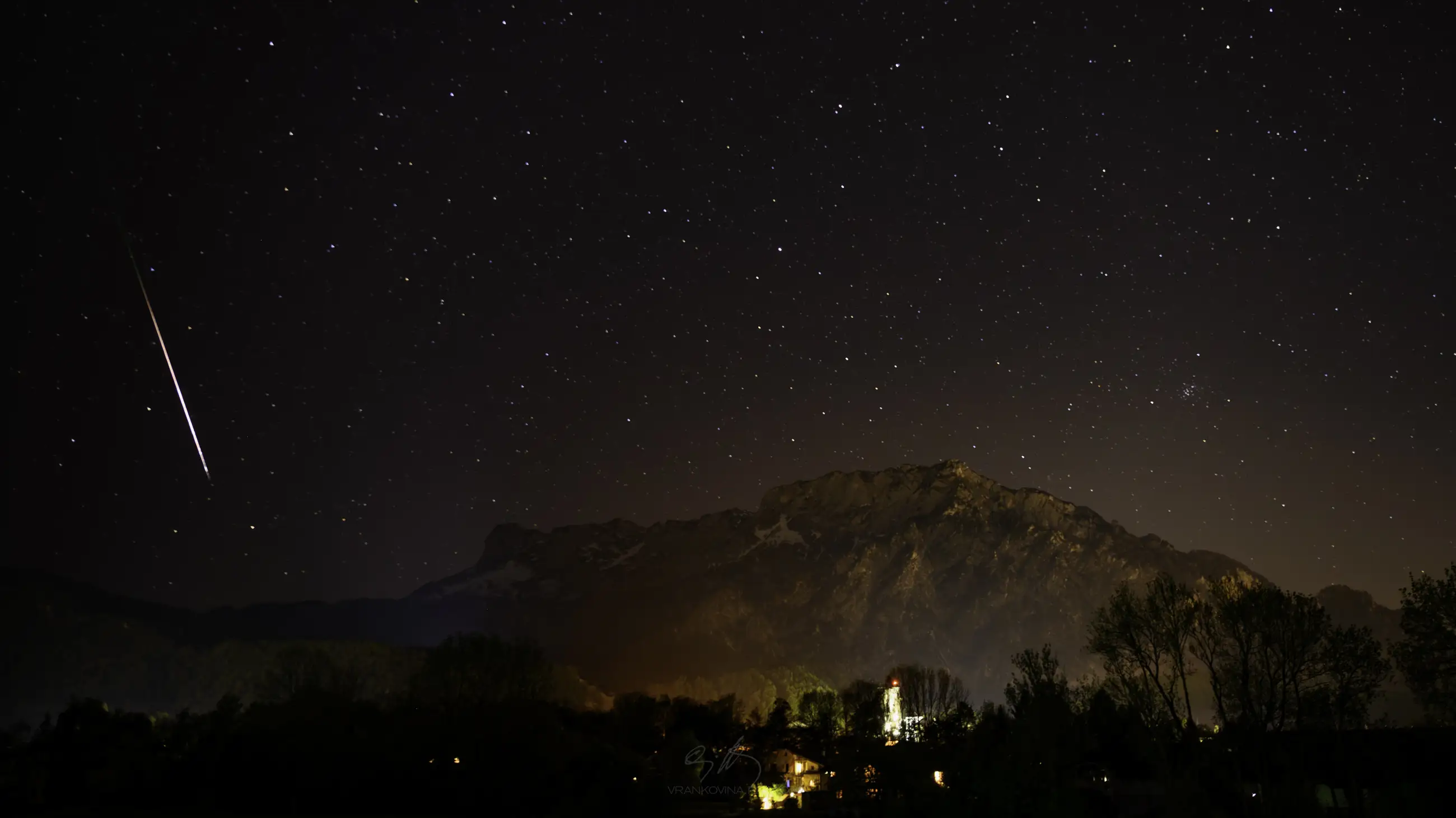 A night sky filled with stars and a shooting star above a mountain range. Below, lights from houses illuminate the landscape, with trees and buildings visible in the foreground.