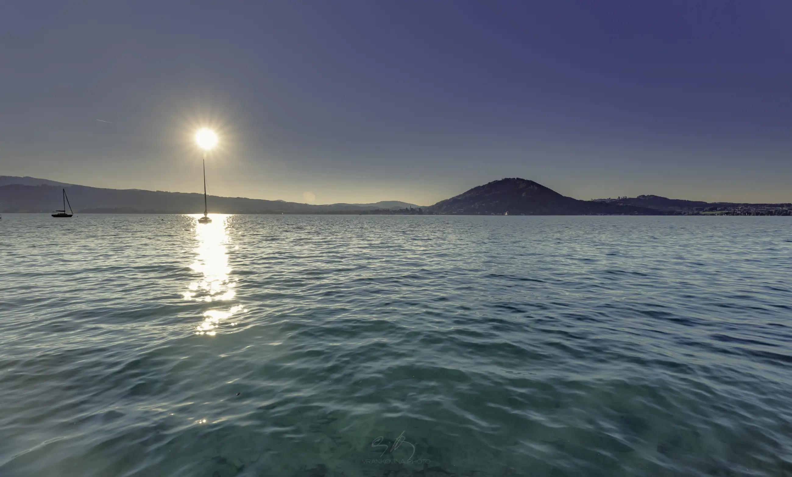 a lake before sunset with a sailing boats anchored at a bouy and small mountain at the opposite shore. turquoise water an deep clear blue sky