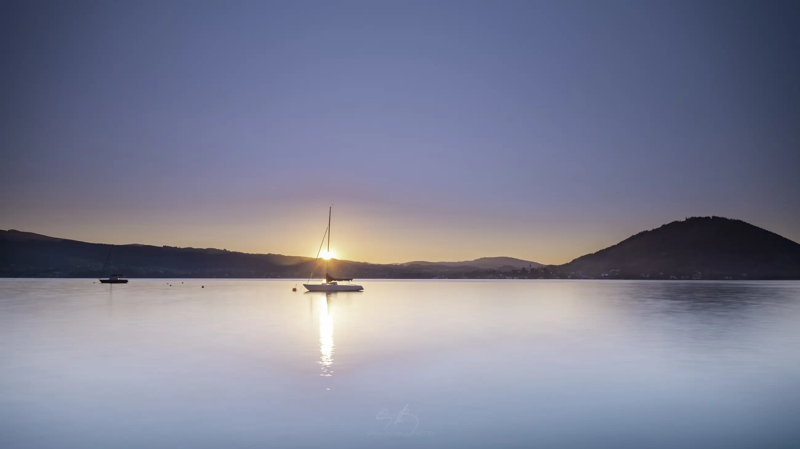 sunset at a lake with a sailing boat in the foreground and small mountain at the opposite shore. Sun is setting exactly behind the mast of the sailingboat