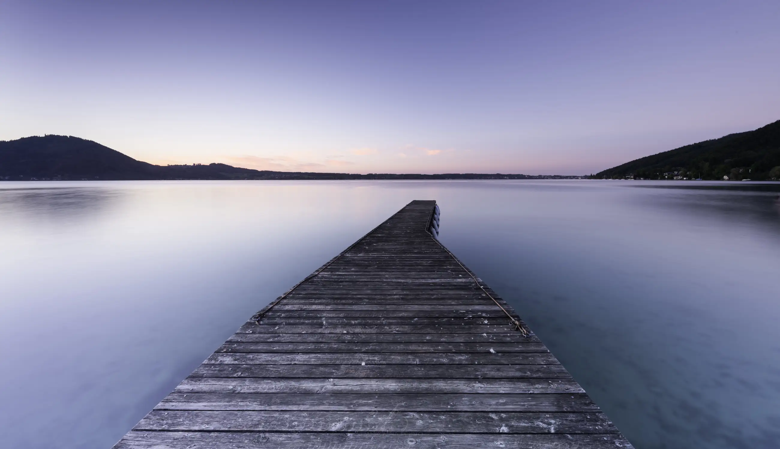 a lake with small mountains at the opposite shore and a jetty in the center after sunset
