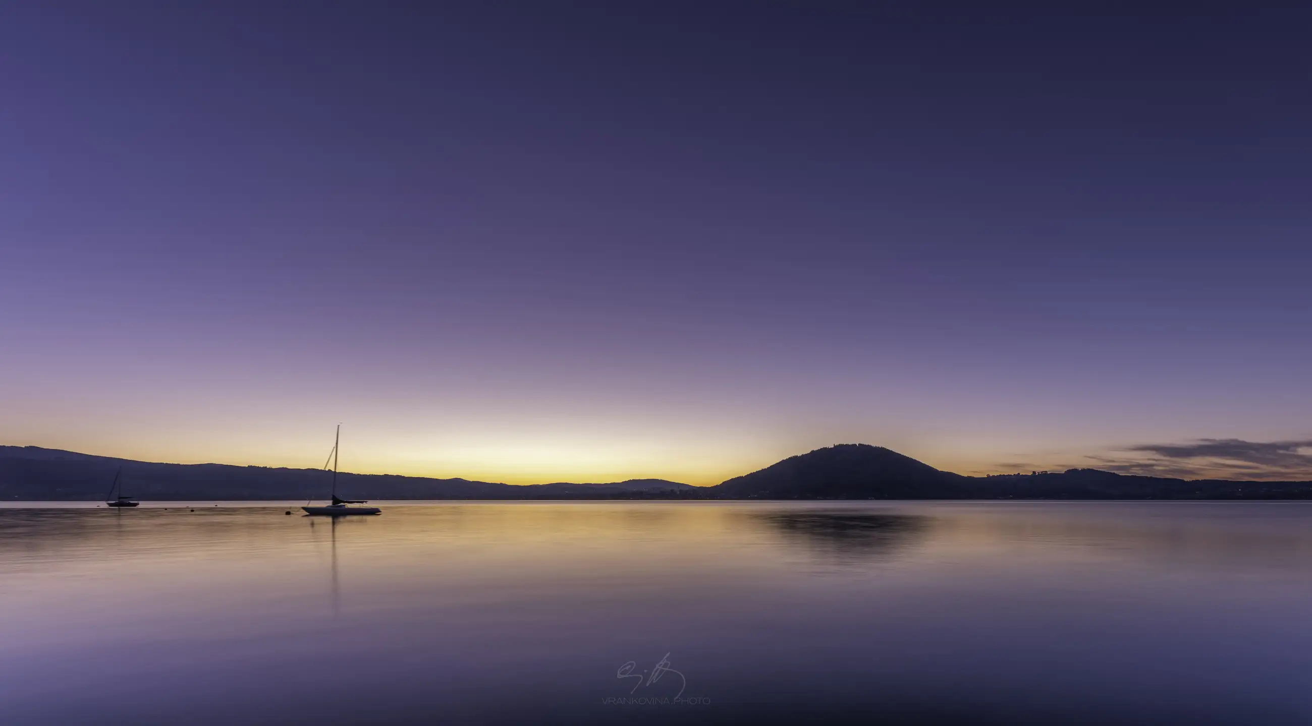 a lake after sunset with a sailing boat in the foreground and small mountain at the opposite shore. Deep violet clear sky