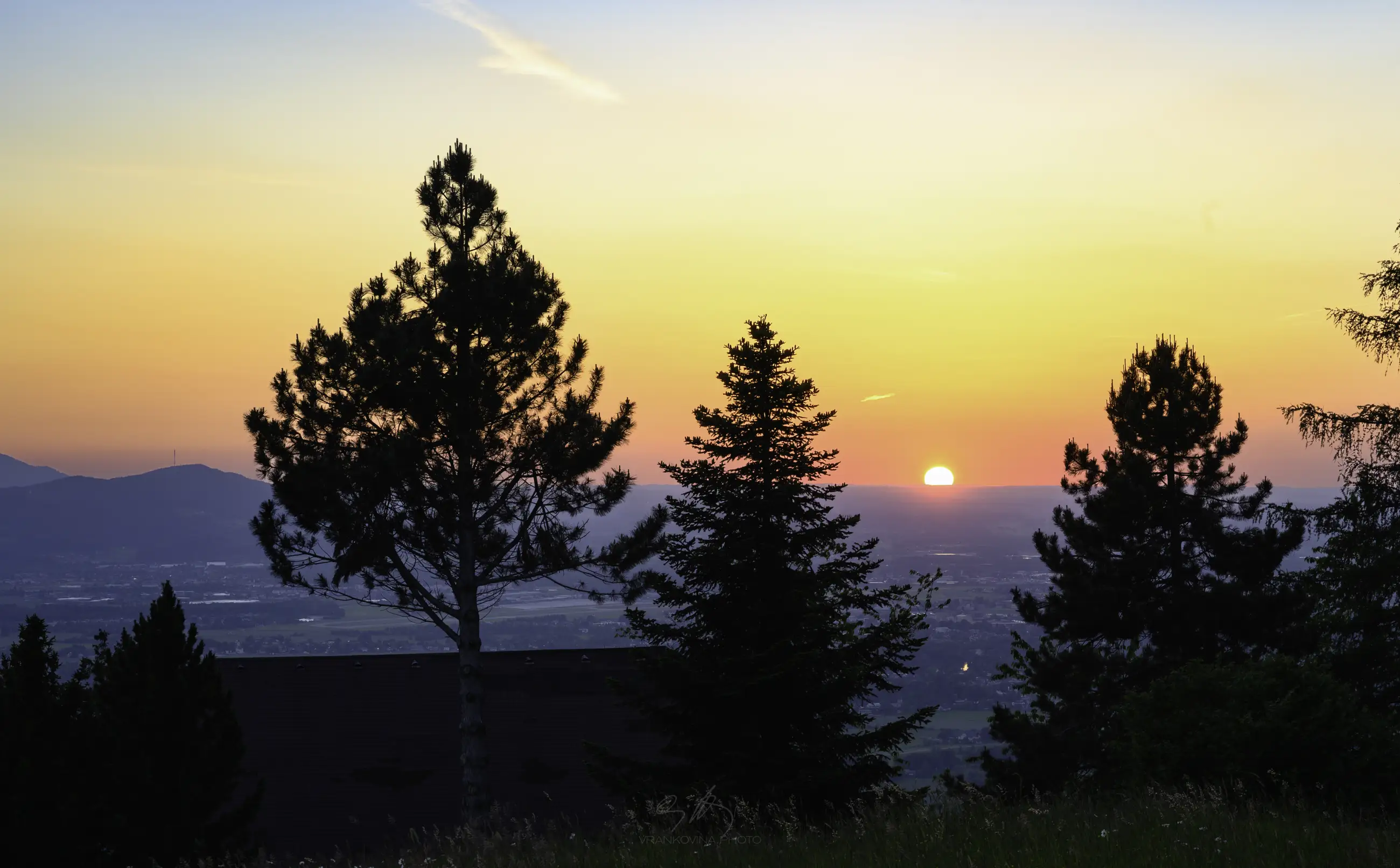 Trees and a house roof in the front, a valley with a sunset in the background, very colorful orange and red sky