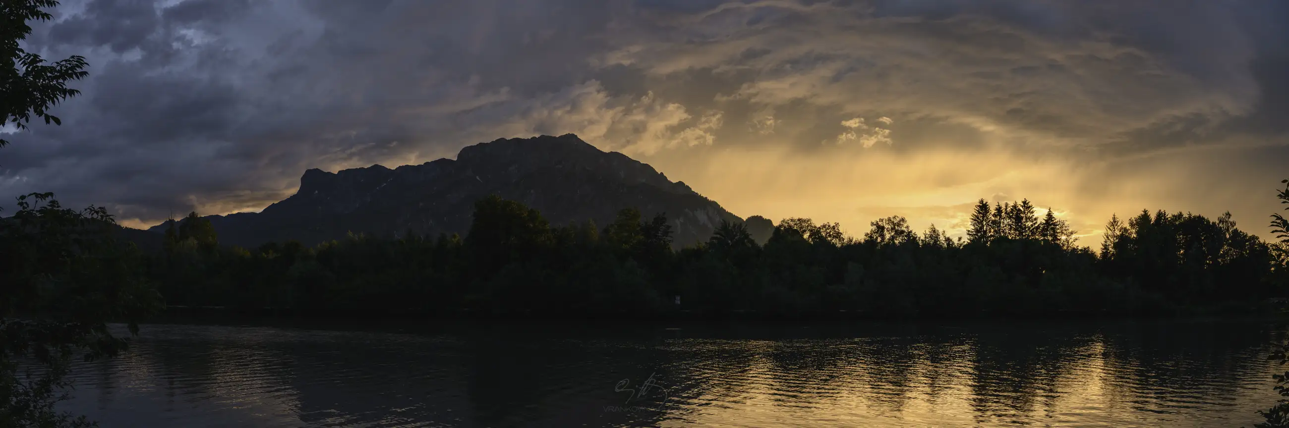 mountain panorama with a river in the foreground and trees on the shore, stormy clouds at sunset with bright orange color
