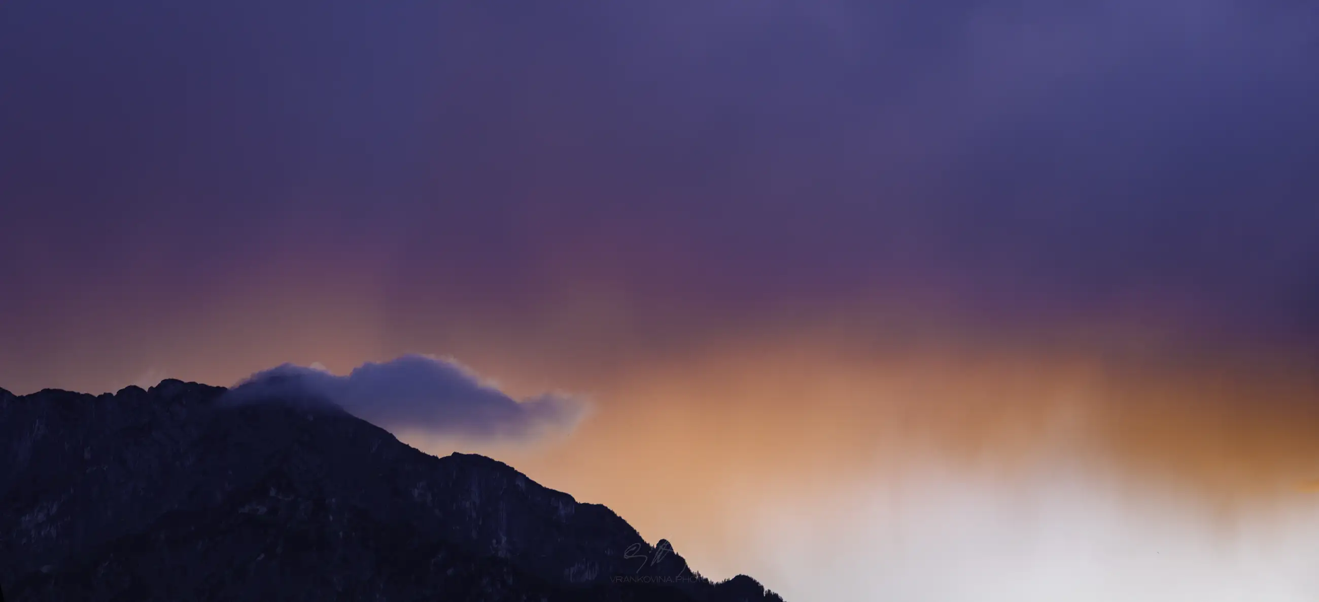 Colorful orange, violet and dark blue sky over a mountain silhouette after storm, a white cloud is sticking to the mountain top
