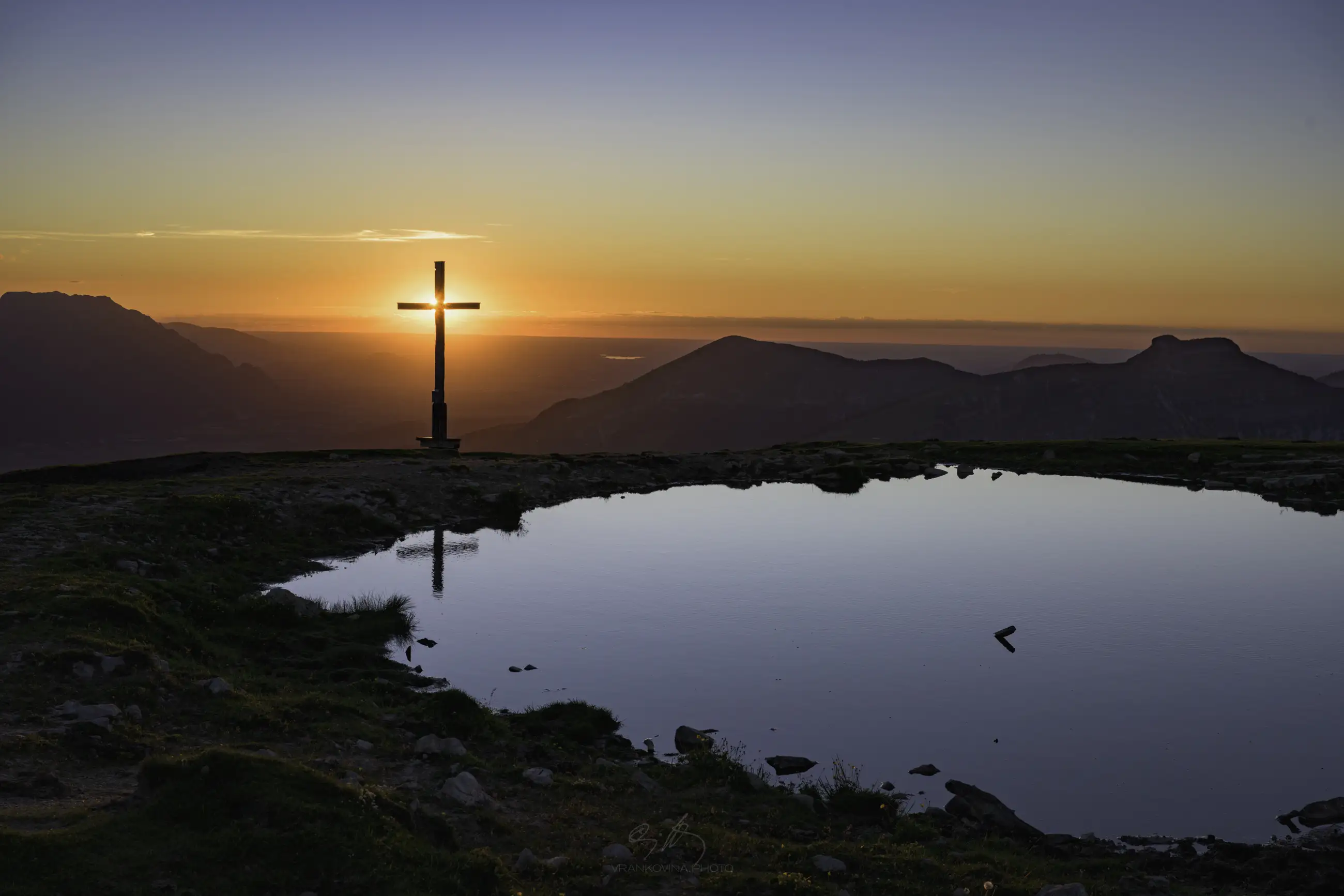 Sunset seen from a mountain, pond and a cross in the foreground, sun is exactly behind the cross