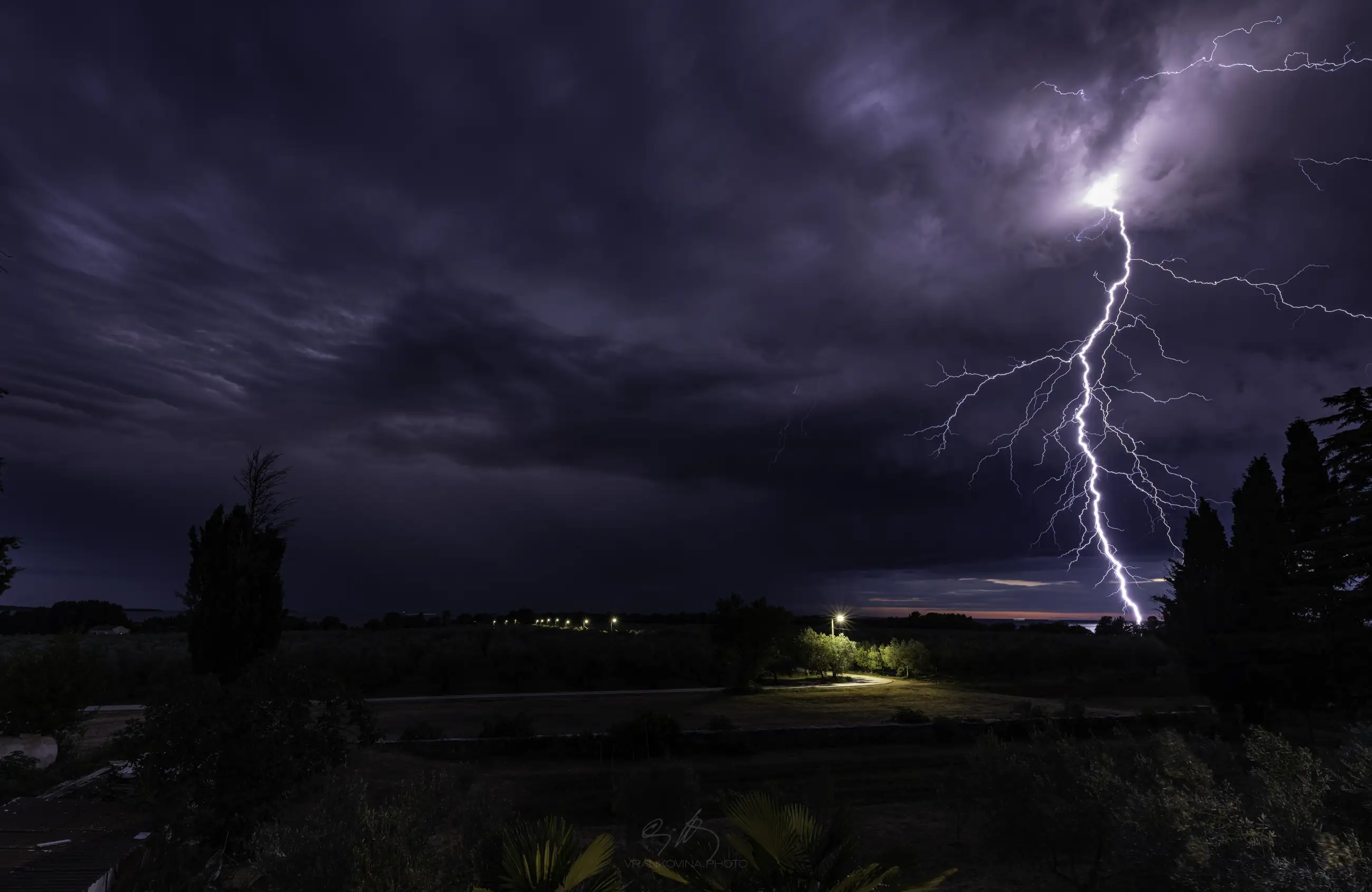 Lightning strikes down from dark storm clouds at night, illuminating the sky above a rural landscape with trees, a field, and a row of distant lights along a road.