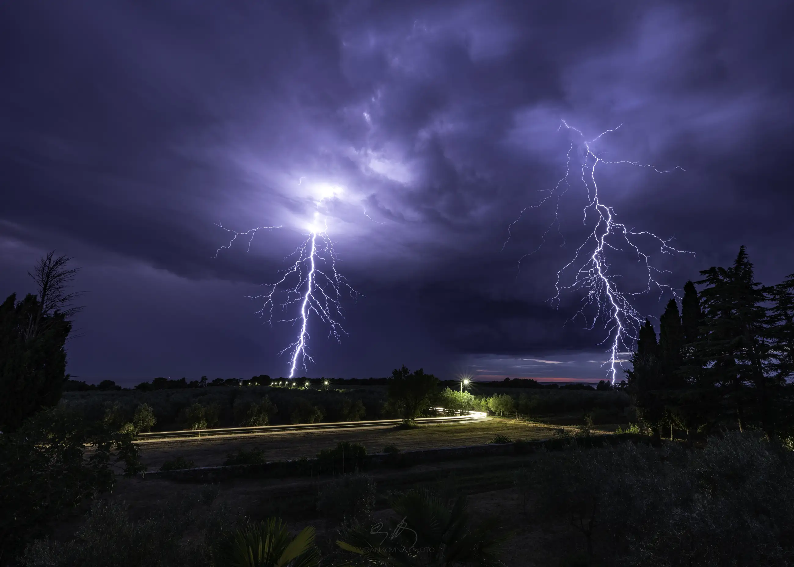 Lightning strikes down from dark storm clouds at night, illuminating the sky above a rural landscape with trees, a field, and a row of distant lights along a road.