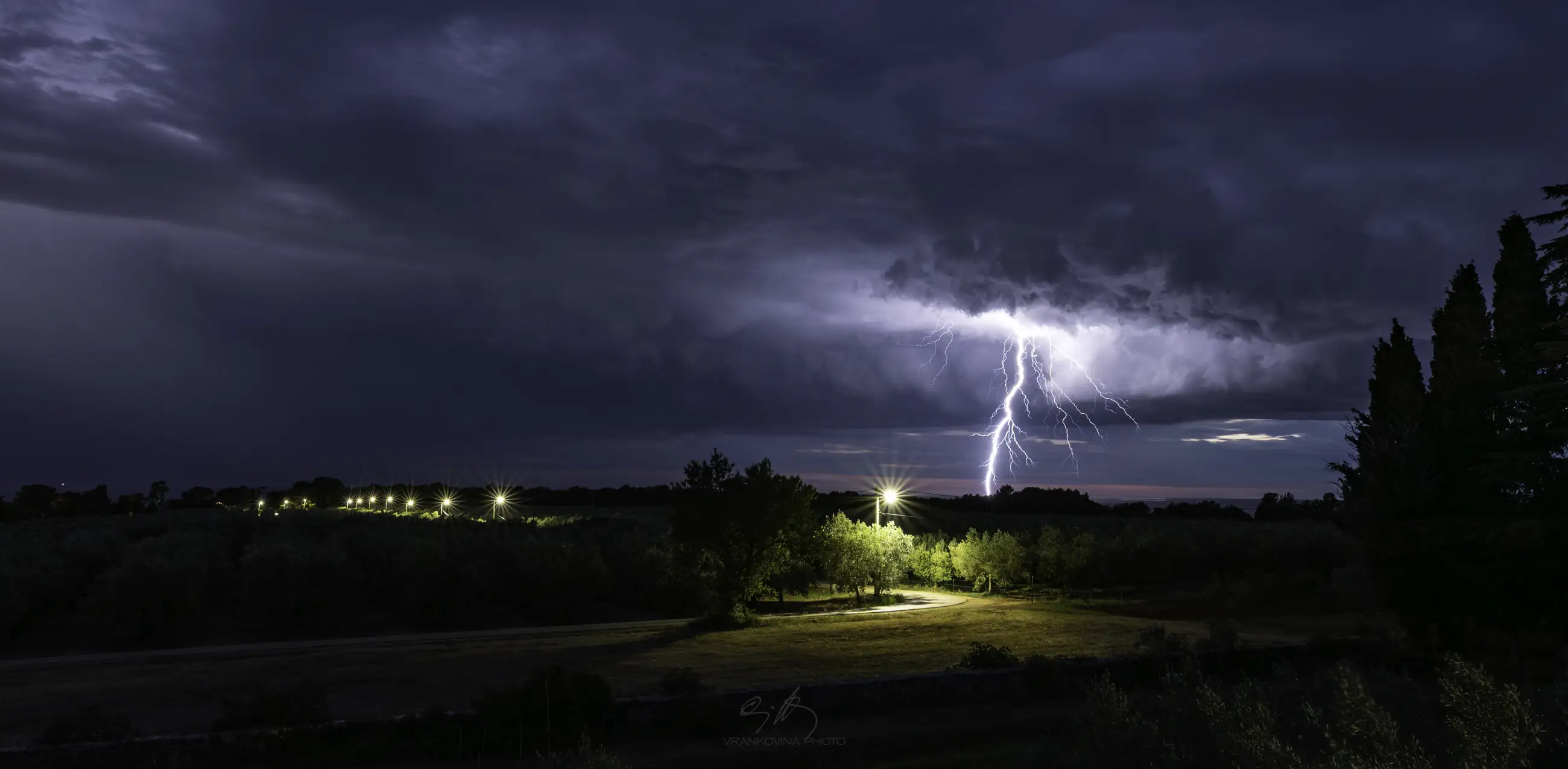 Lightning strikes down from dark storm clouds at night, illuminating the sky above a rural landscape with trees, a field, and a row of distant lights along a road.