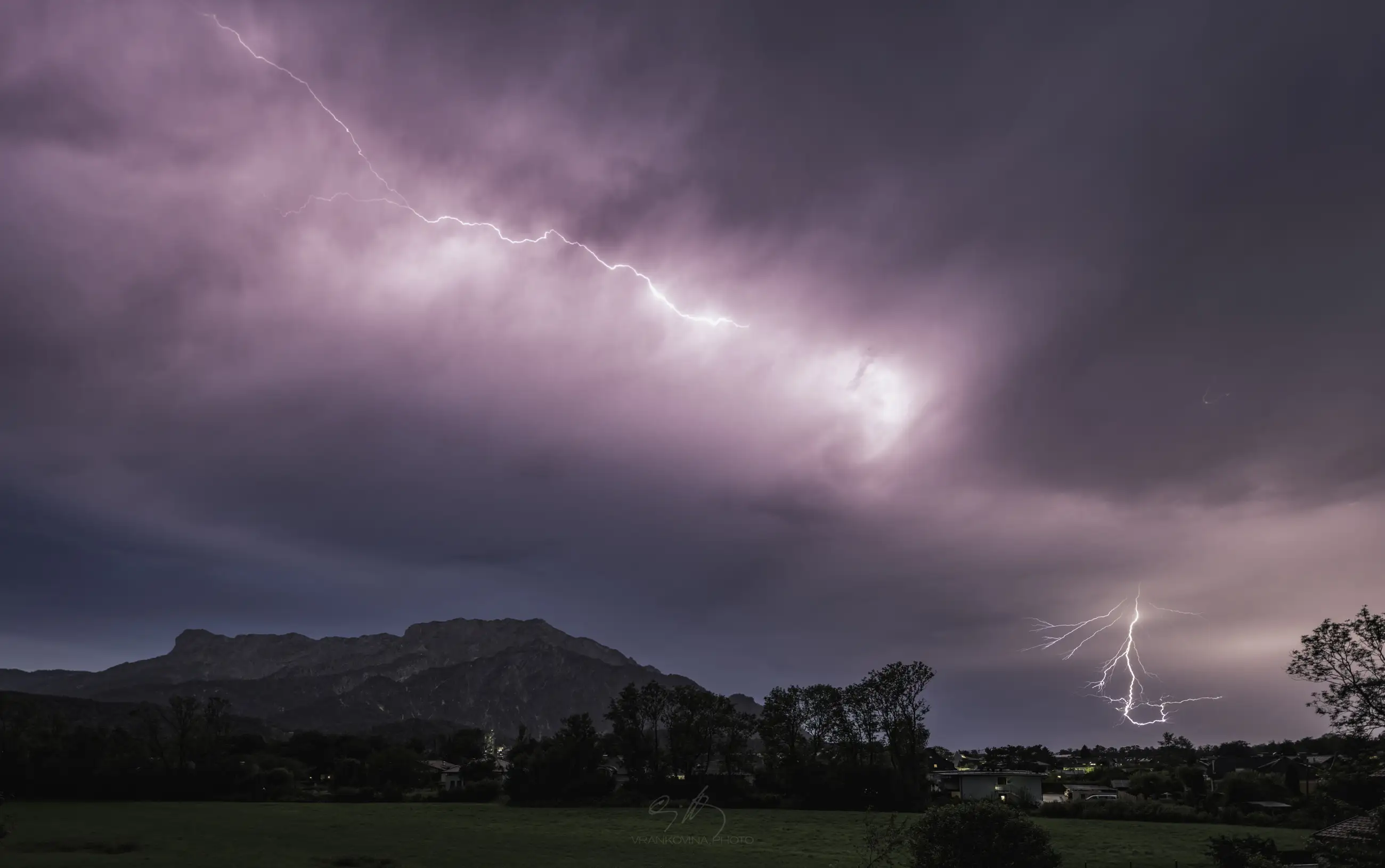 A vivid bolt of lightning streaks across a dramatic, cloudy sky over a dark mountain and a small town, illuminating the landscape and grassy field below.