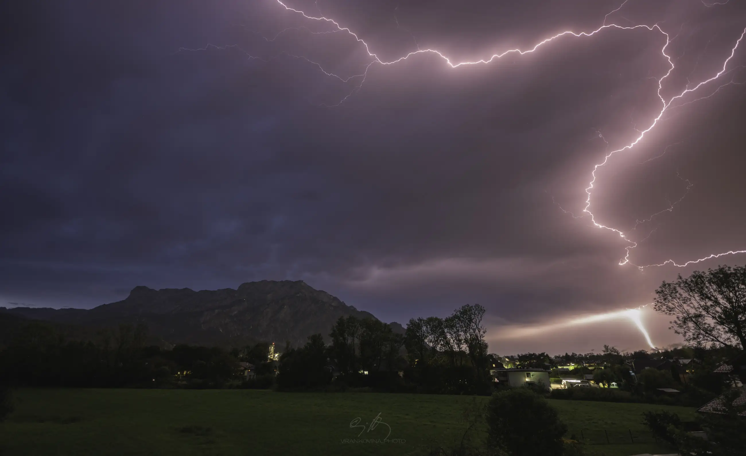 A vivid bolt of lightning streaks across a dramatic, cloudy sky over a dark mountain and a small town, illuminating the landscape and grassy field below.