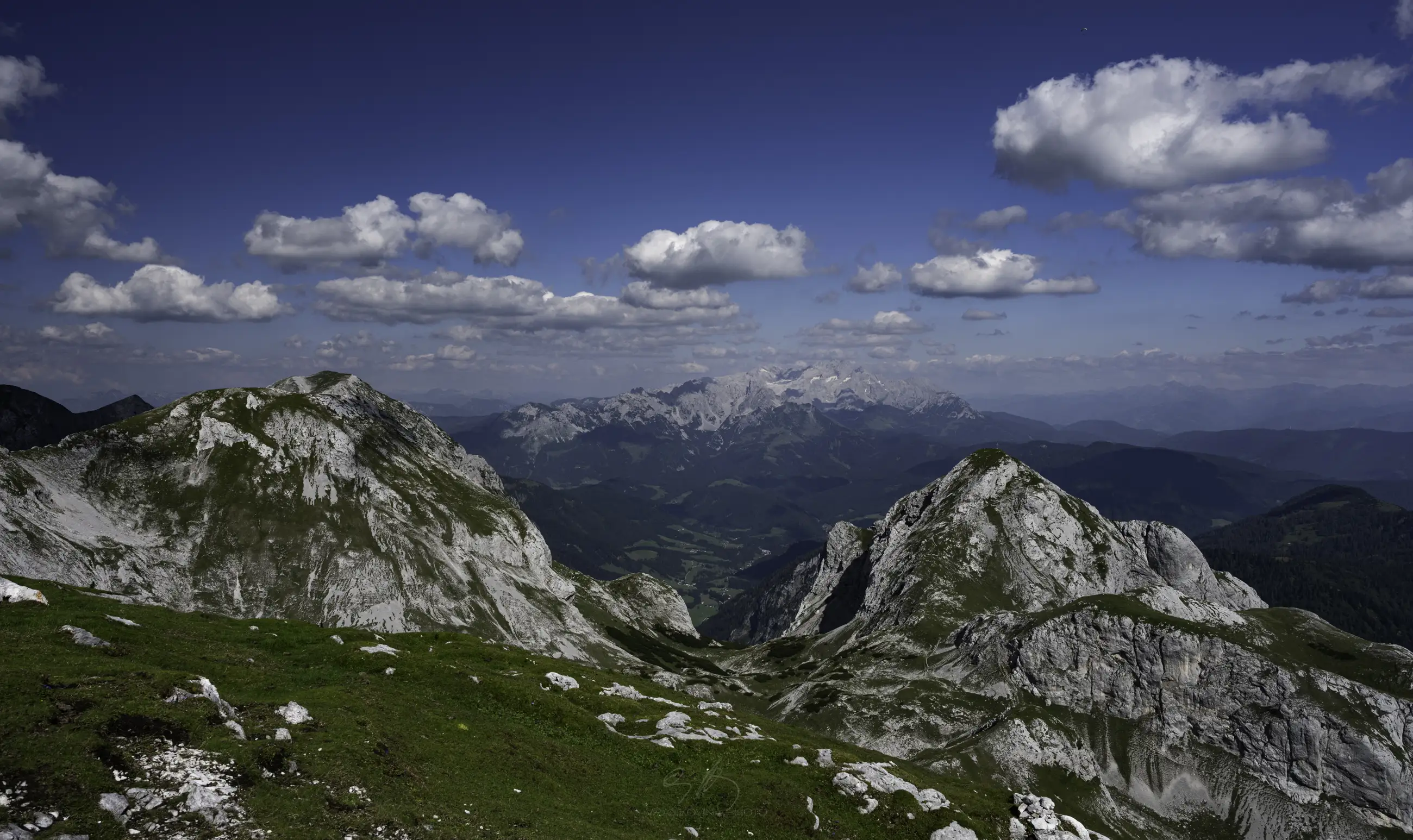 Rocky mountain peaks with patches of green grass in the foreground, under a deep blue sky dotted with scattered white clouds. Distant mountains and valleys are visible under the clear weather.