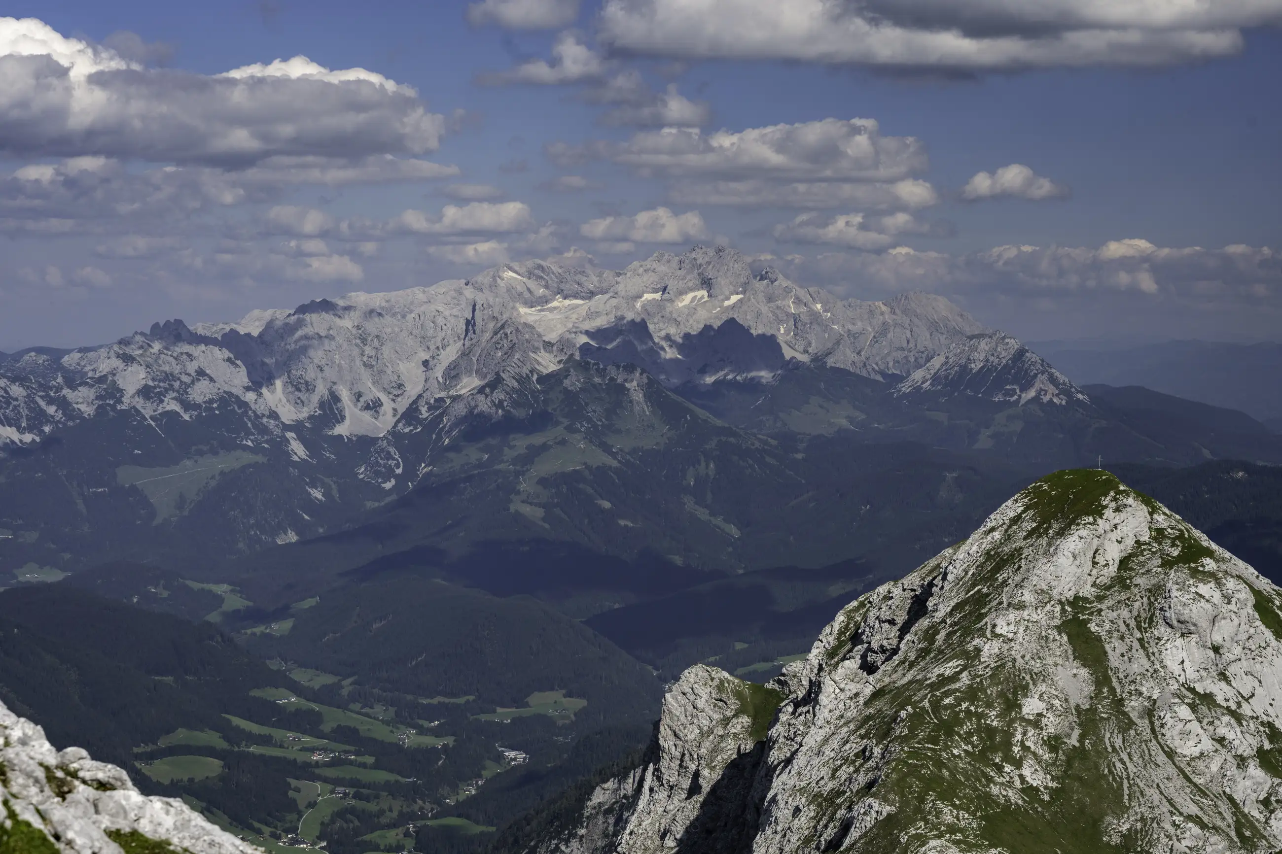 A panoramic view of alpine mountains with rocky peaks, green valleys, and scattered clouds in a blue sky. Sunlight highlights patches of grass and trees on the slopes.